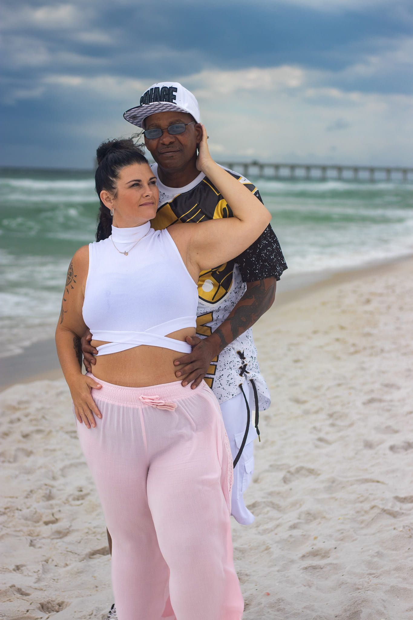 Couple posing together on sunny beach with pier visible in background.