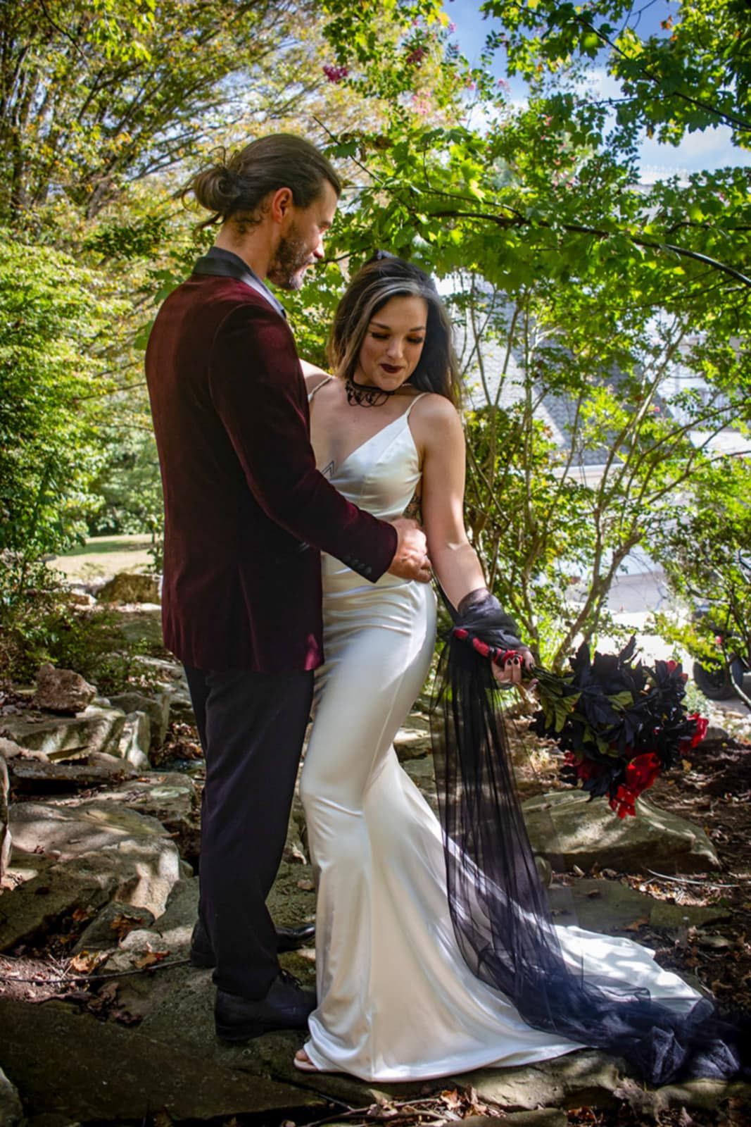 A couple in formal attire standing on stone steps surrounded by lush greenery.