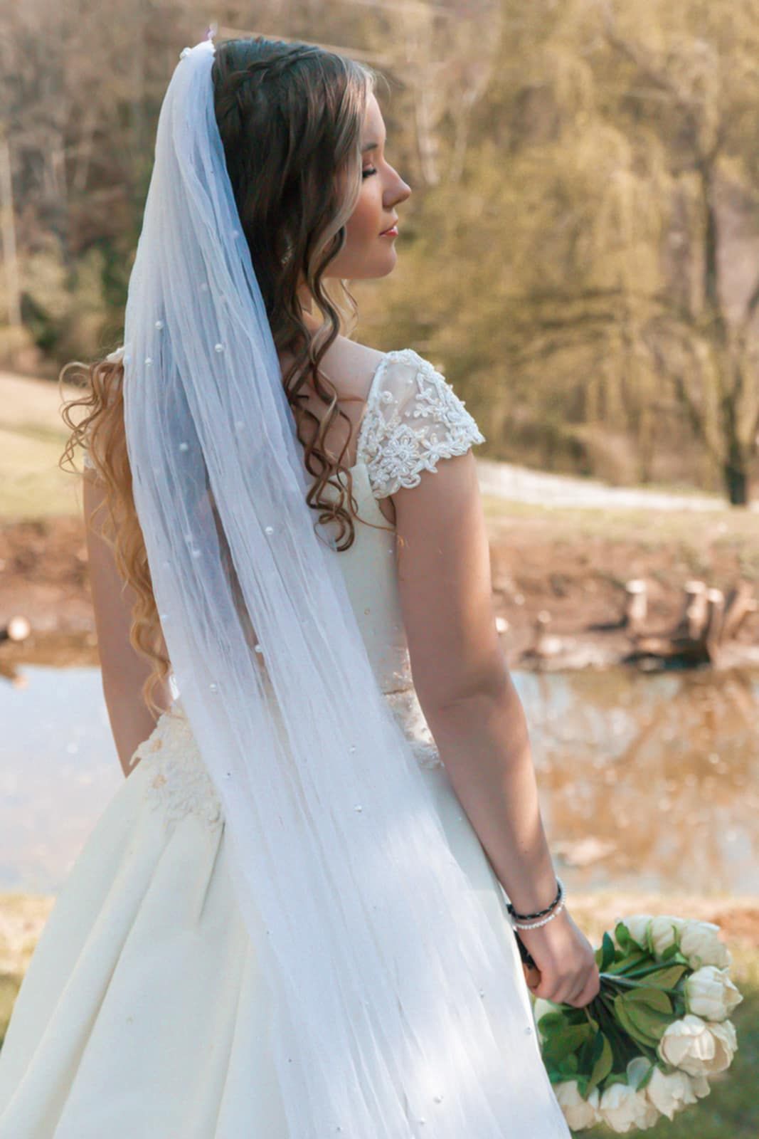 A side profile of a bride in a lace cap sleeve gown with flowing veil against an autumn background.