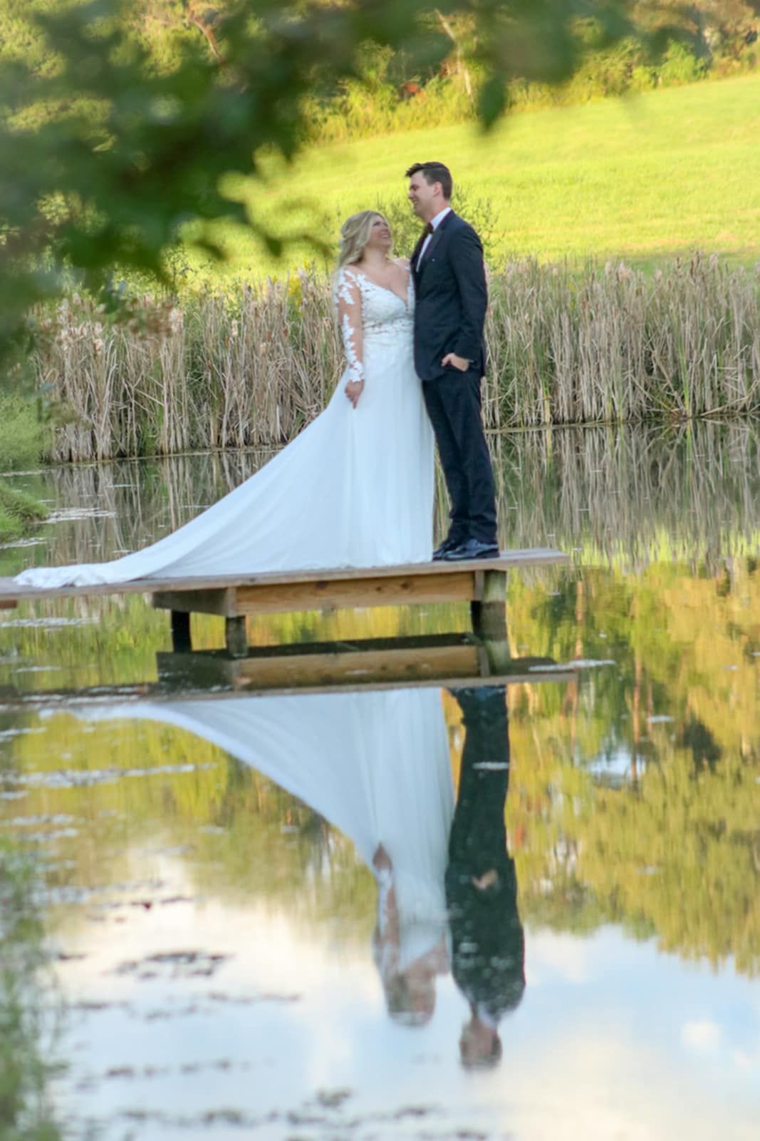 A wedding couple poses on a wooden dock with their reflection mirrored perfectly in the still water below.