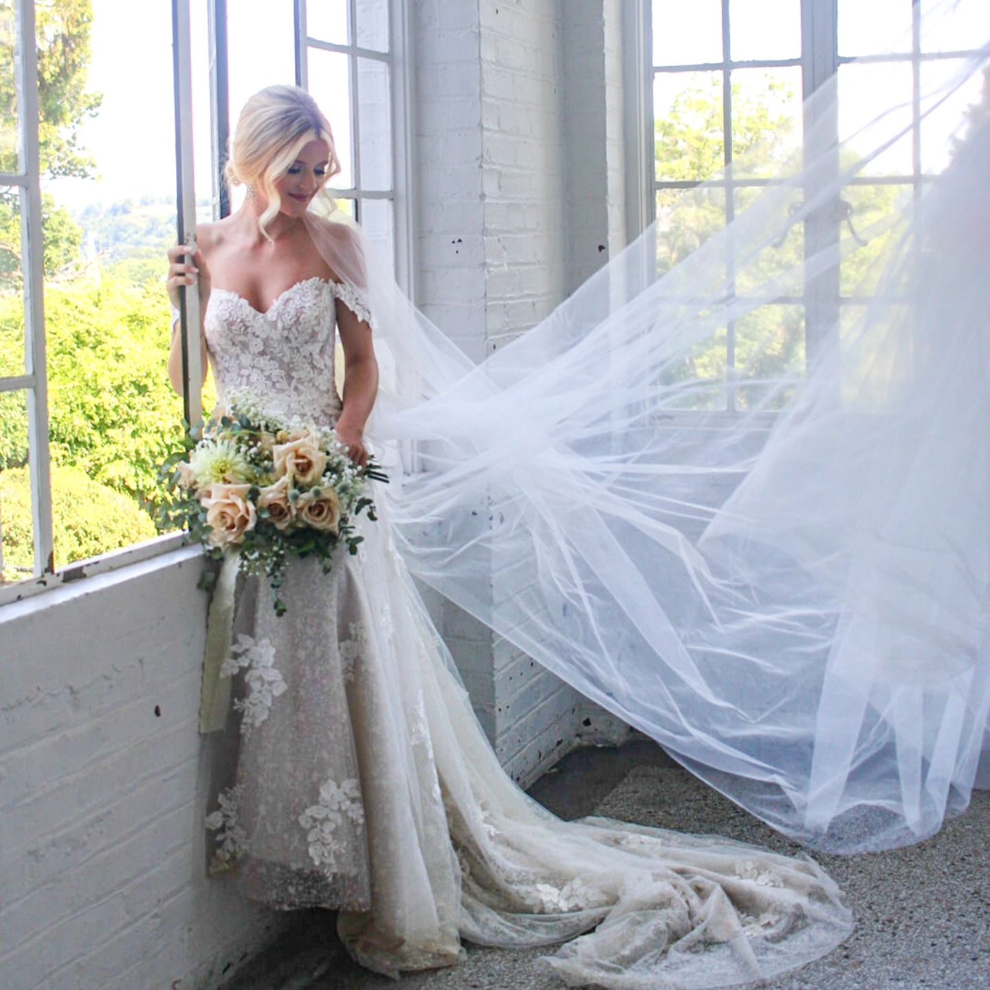Bride in lace gown poses by window with flowing veil and pastel floral bouquet.