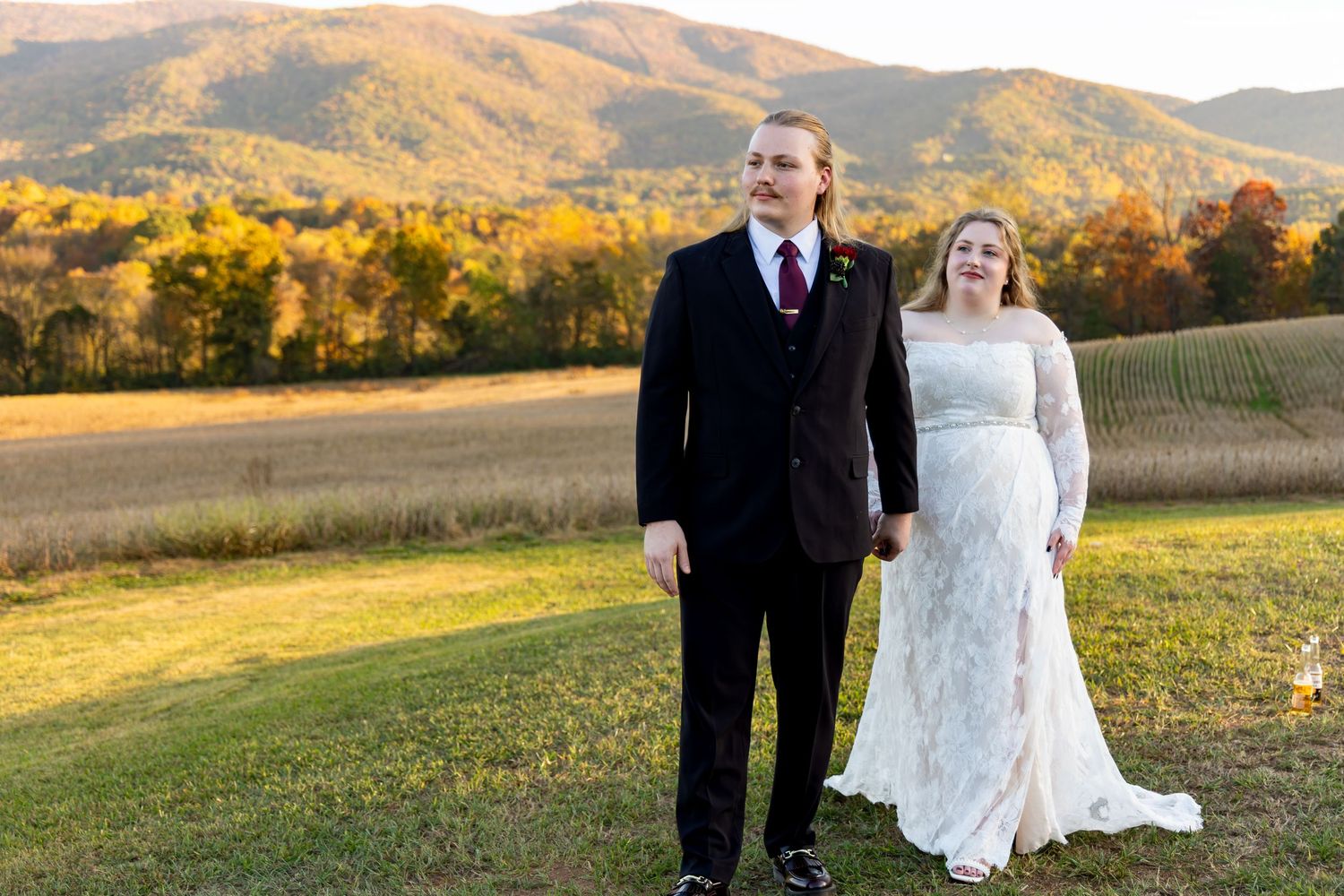 A newlywed couple stands in autumn sunlight on a hillside with golden mountains in the background.