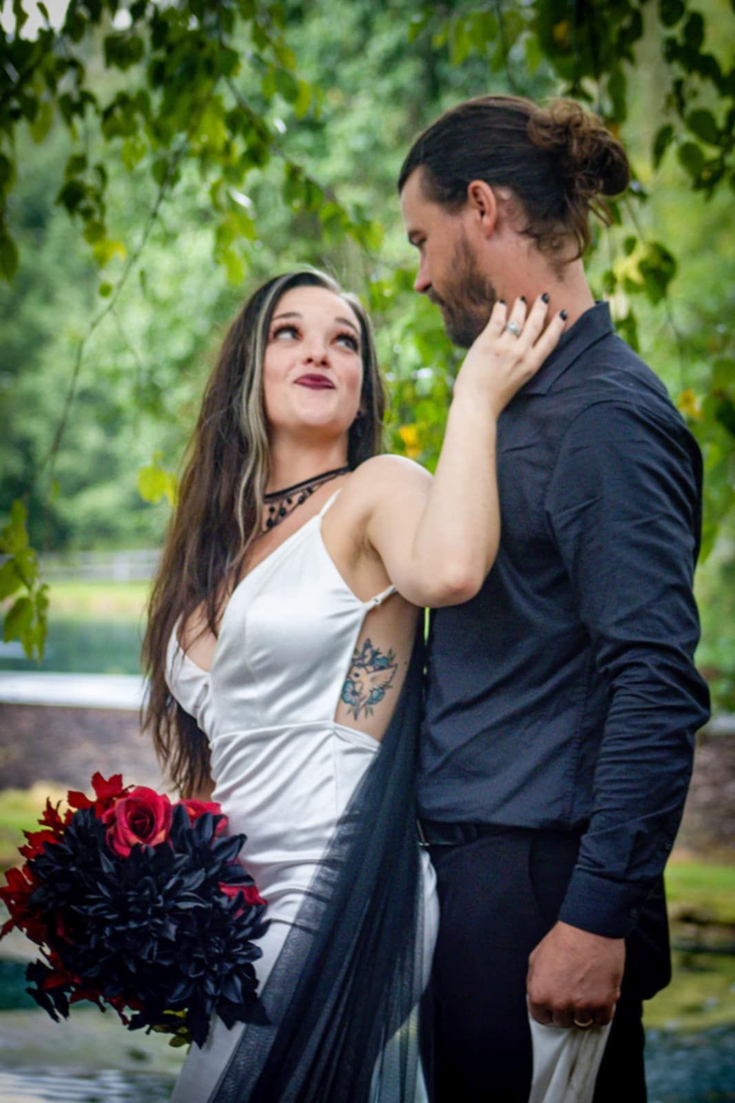 A romantic couple in contrasting black and white attire pose with a dark red bouquet among green foliage.