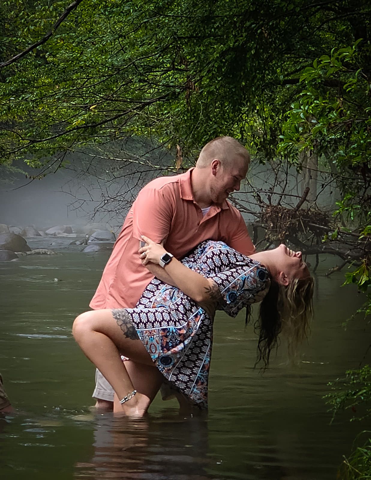 Romantic couple poses for dramatic dip pose in water surrounded by green foliage.