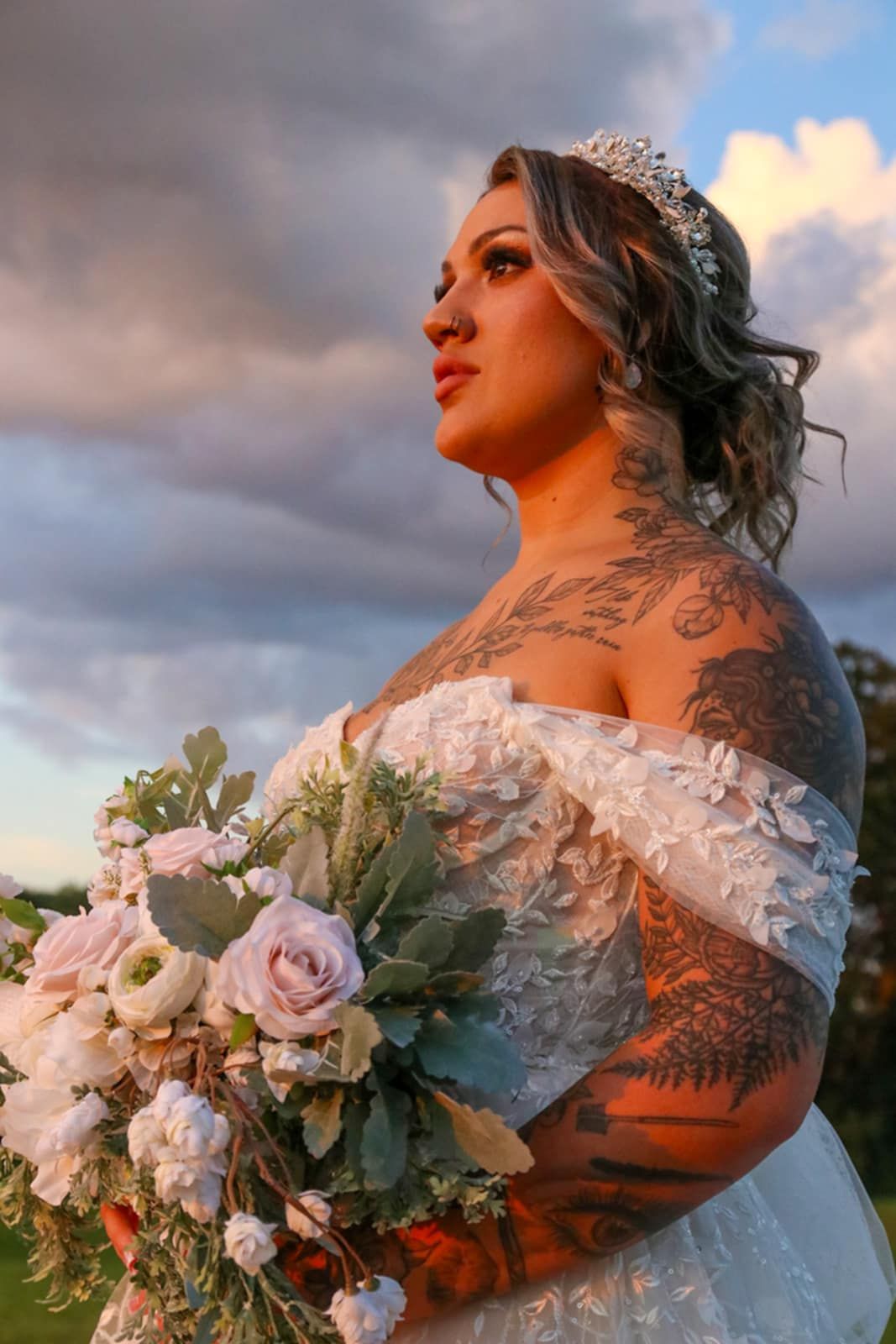 A bride in a white lace dress and crown poses at sunset holding a bouquet of pale pink roses.