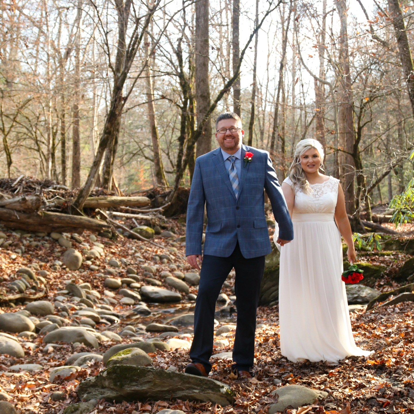 A couple in wedding attire stands together on a rocky forest path covered in autumn leaves.