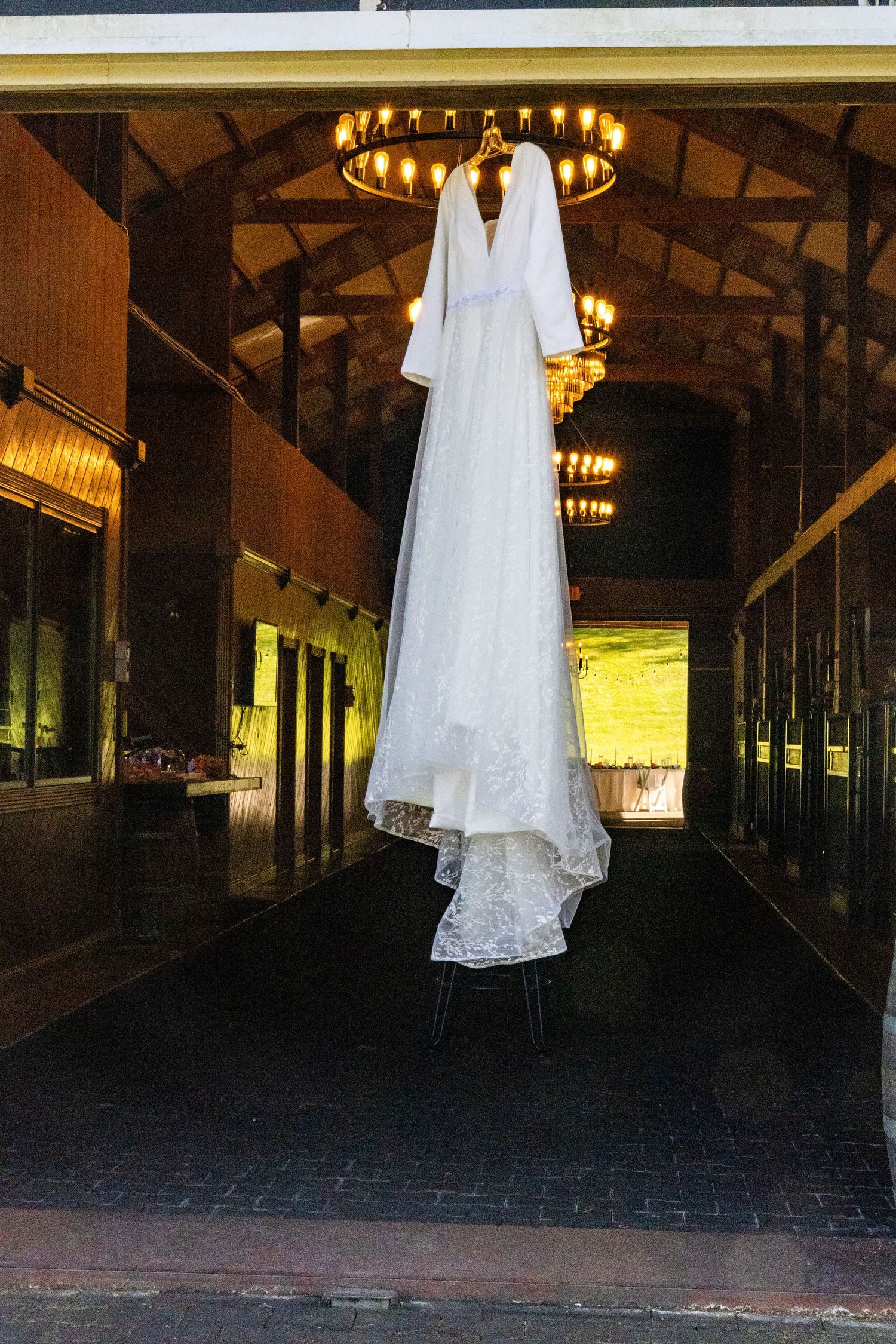 Elegant white wedding dress hanging in a dimly lit rustic wooden hallway with chandeliers.