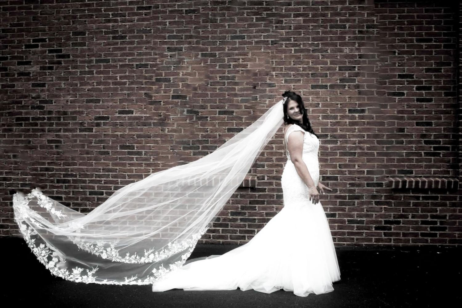 A dramatic black and white photo of a bride with a long flowing veil against a brick wall.