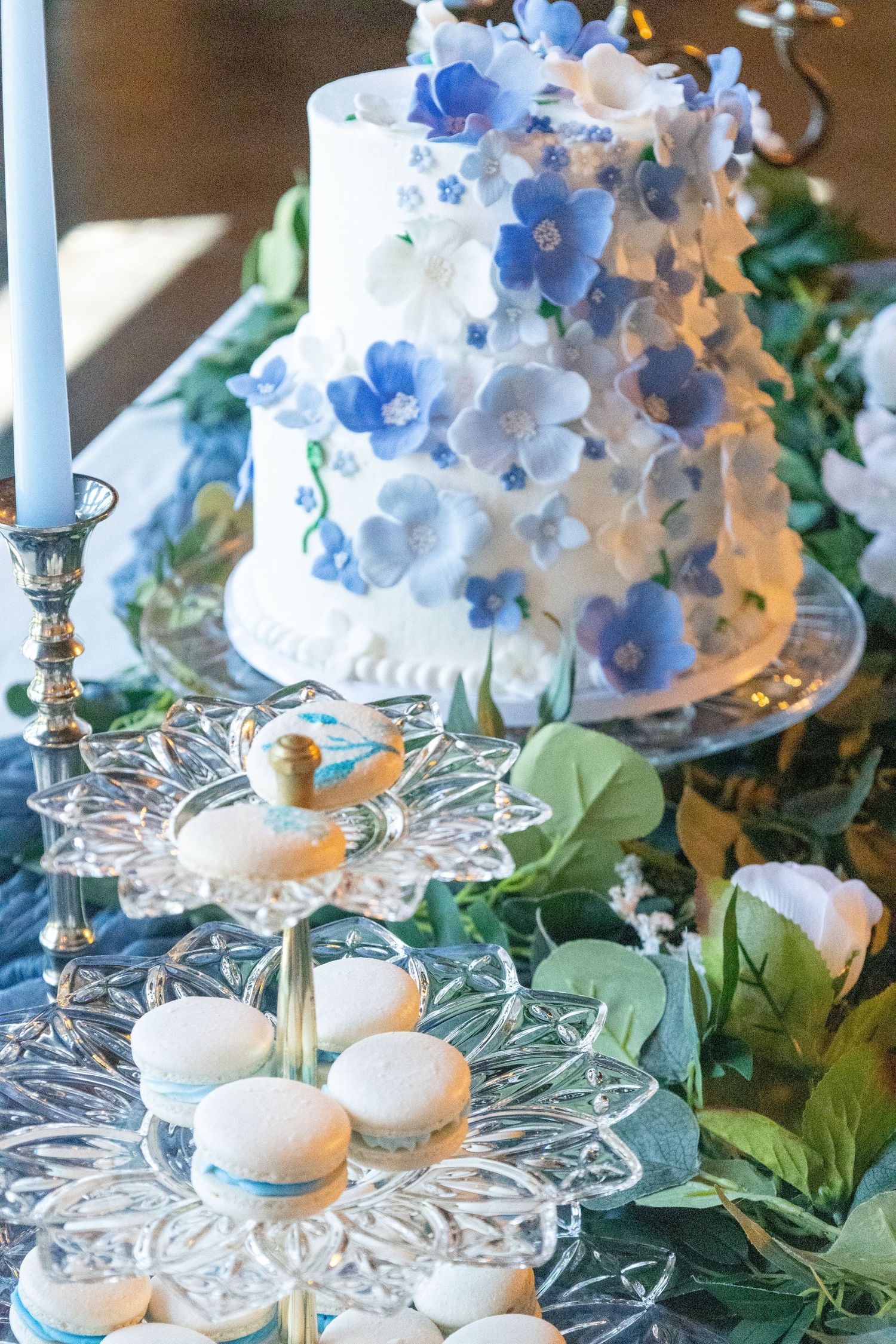 Wedding dessert table featuring a white cake with blue flowers and macarons on crystal stands.