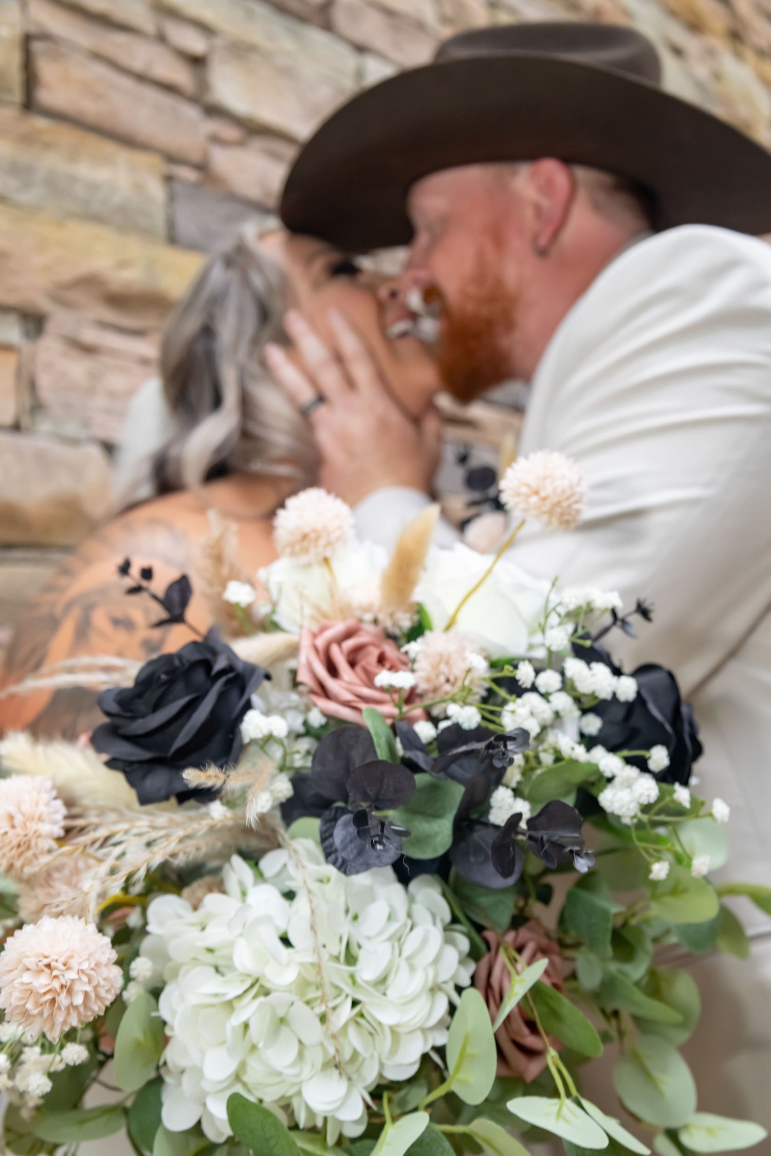 A romantic wedding bouquet with black and white flowers, blush roses, and greenery in front of a stone wall.