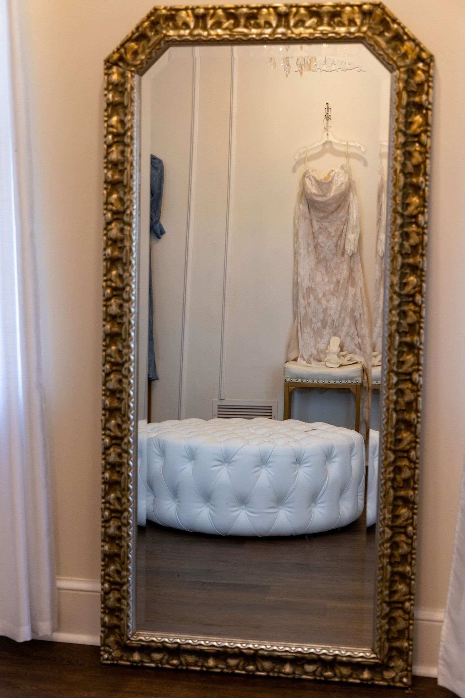 Ornate gold mirror reflecting wedding dress and tufted ottoman in dressing room.