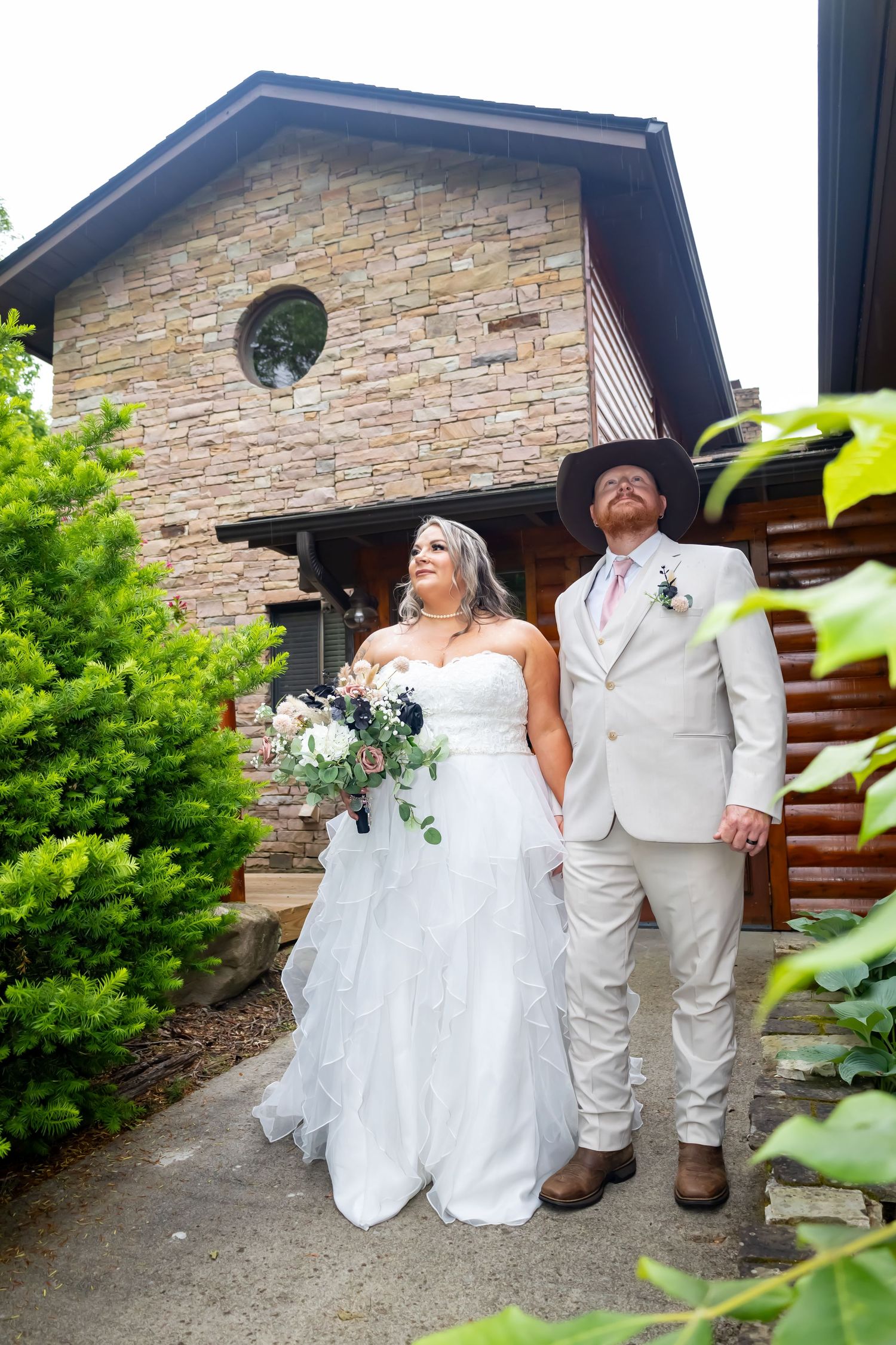 A couple in wedding attire stands outside a stone building with rustic architectural details.