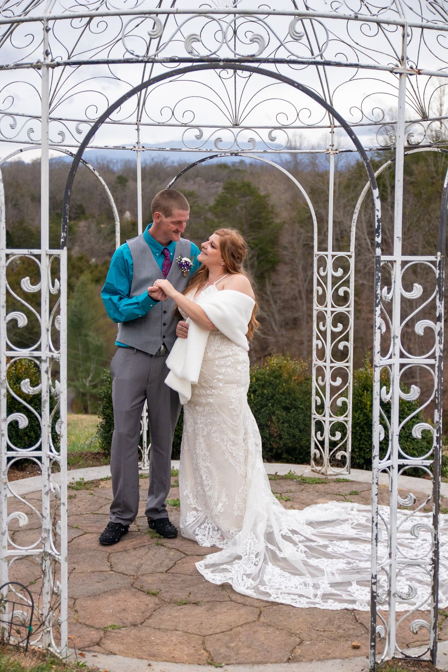 A couple shares a tender moment under an ornate white metal gazebo with decorative scrollwork during their outdoor wedding ceremony.