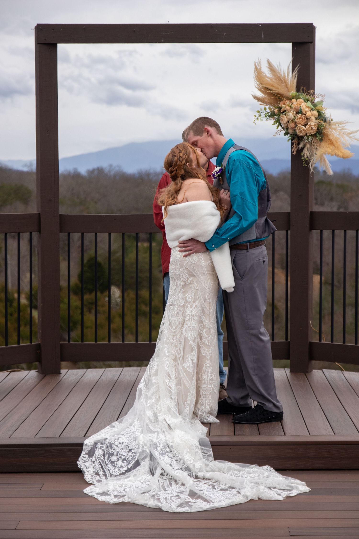 A romantic wedding couple embrace on a rustic wooden deck overlooking scenic mountain views with dried floral decor.