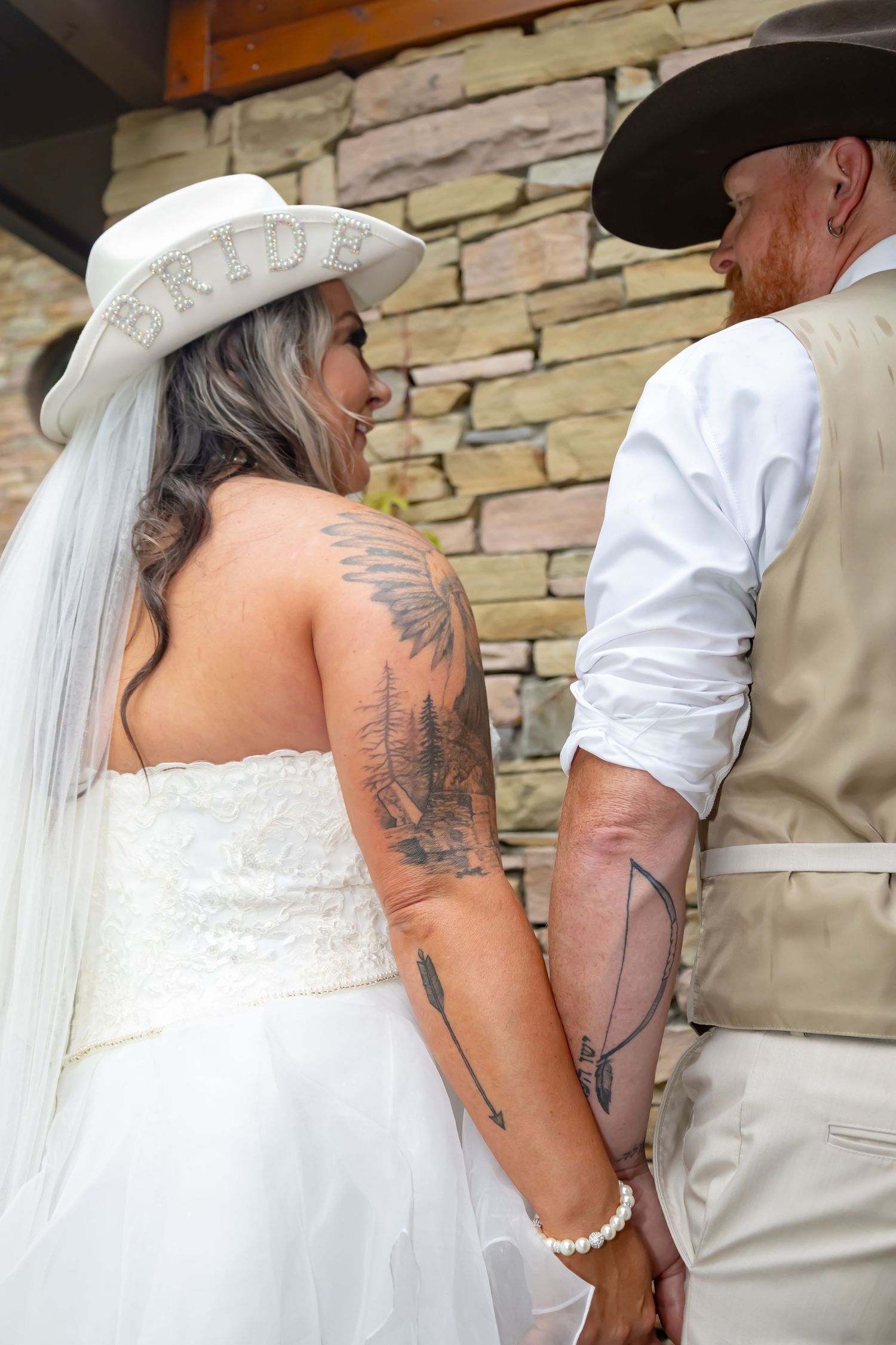 A bride in a white dress and cowboy hat holds hands with her groom in western attire against a stone wall.