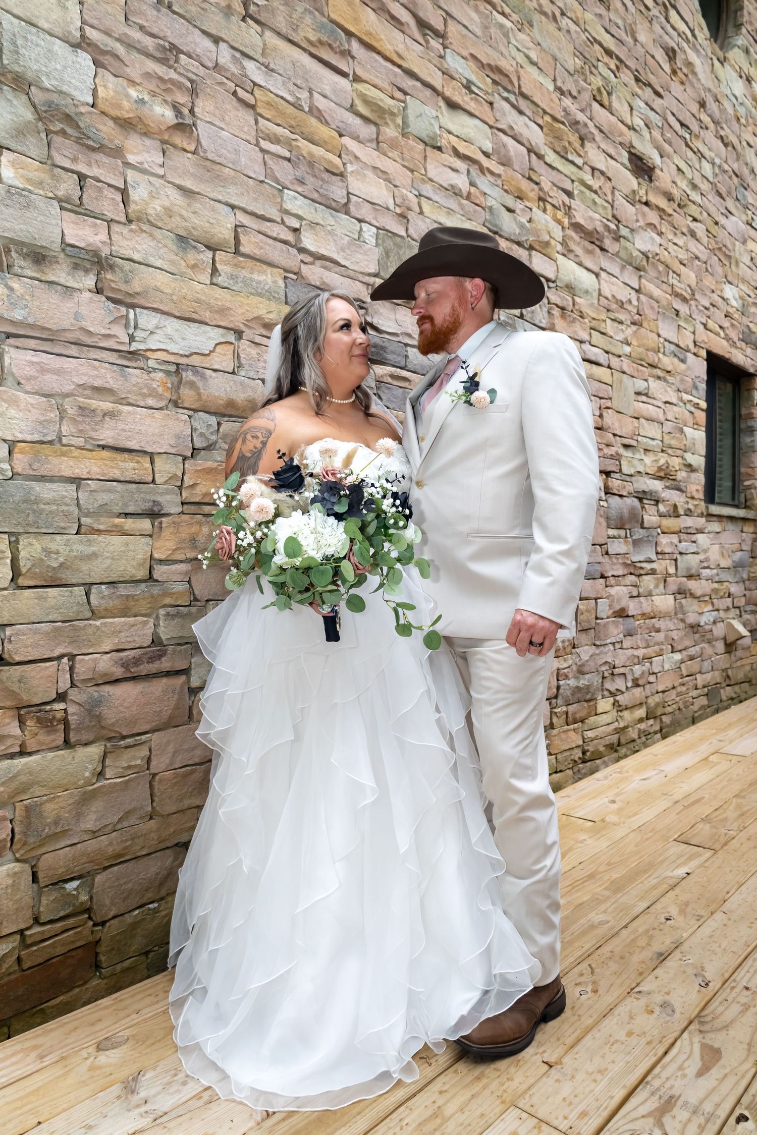 A couple shares a romantic moment against a rustic stone wall, with the bride in white and groom wearing a matching suit and cowboy hat.