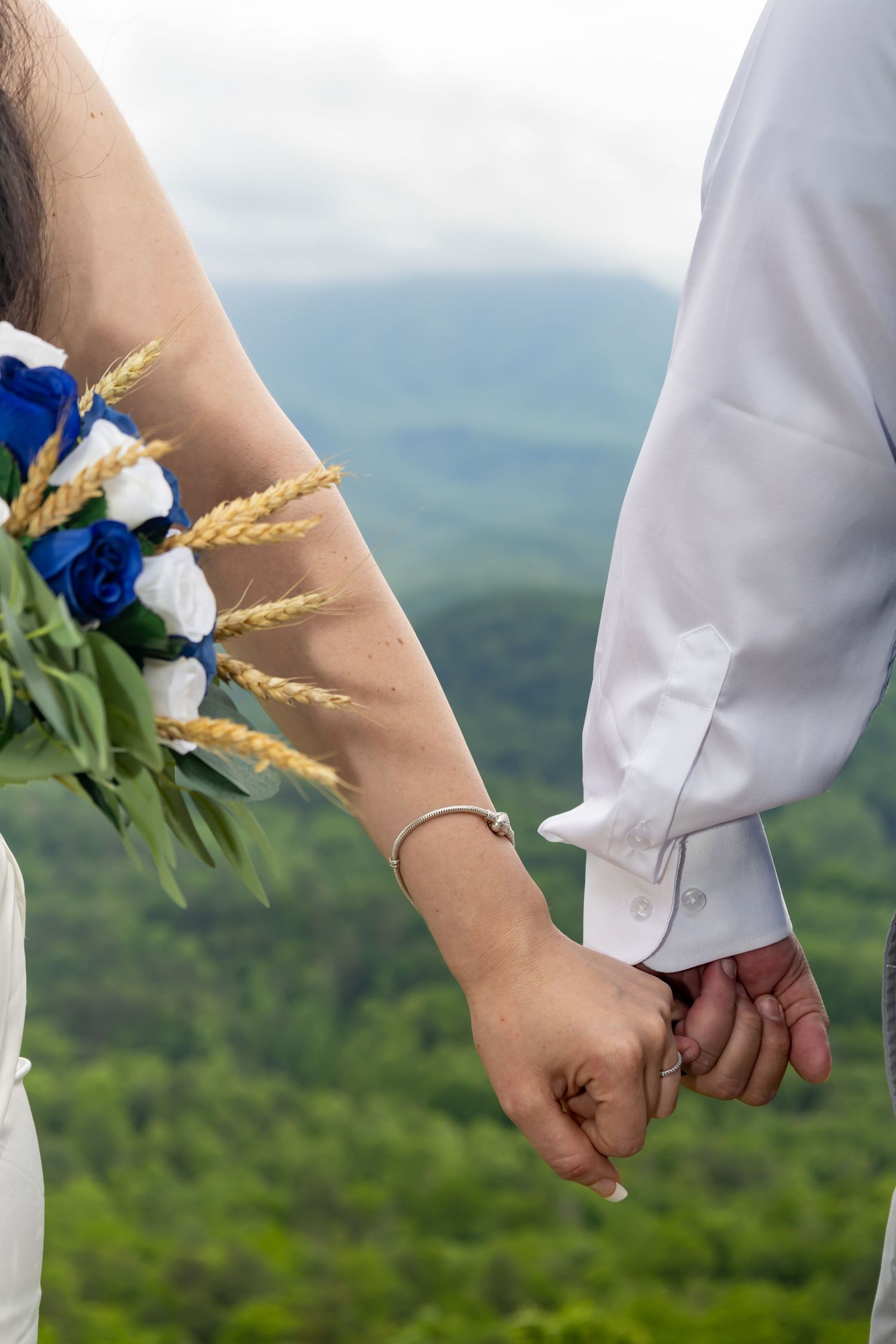 Close-up of newlyweds holding hands with blue floral bouquet against mountain landscape.