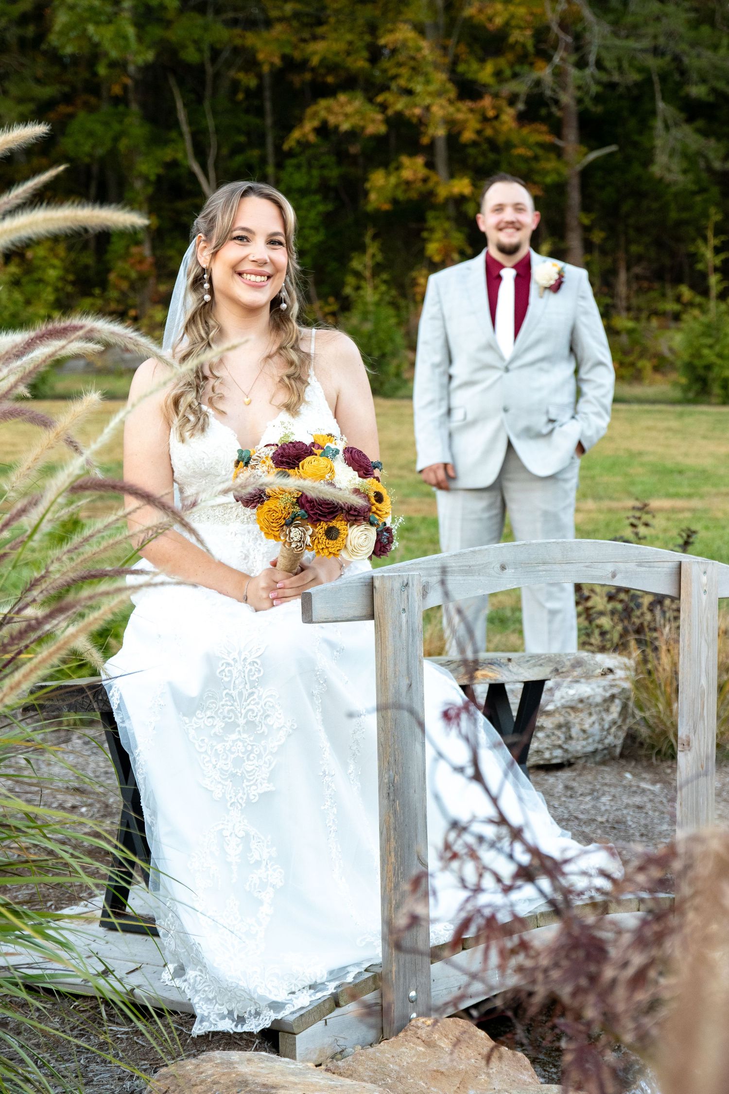 Bride in white dress holds autumn bouquet on wooden bridge while groom in light suit stands behind her.