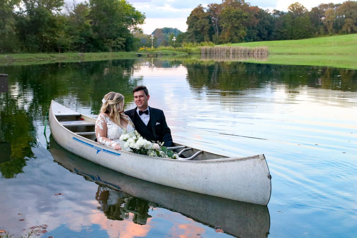 A couple shares a romantic moment in a white canoe on a serene lake surrounded by trees and green landscape.
