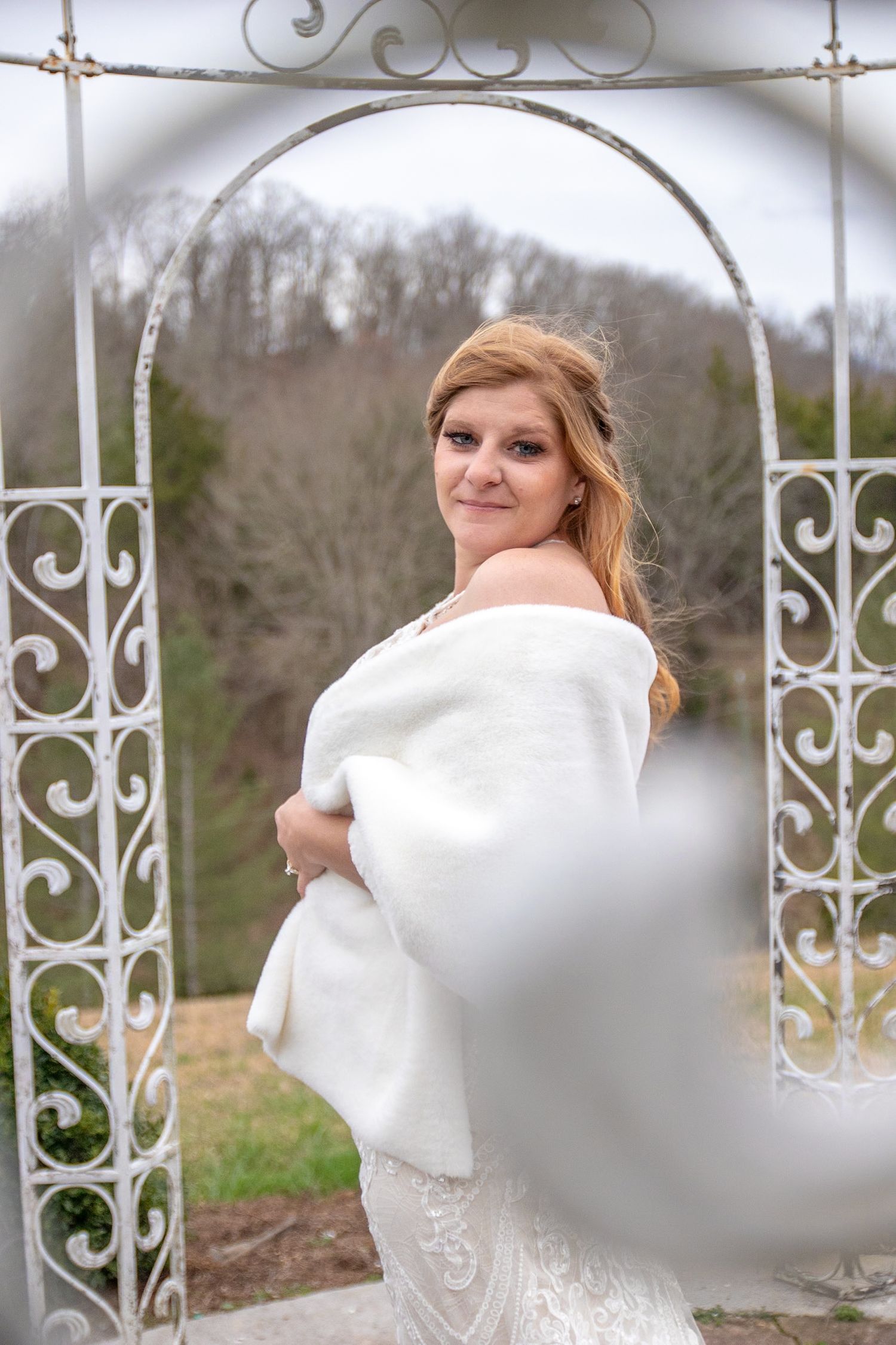 Elegant bride wearing off-shoulder gown poses in front of white metal garden arch during outdoor portrait session.