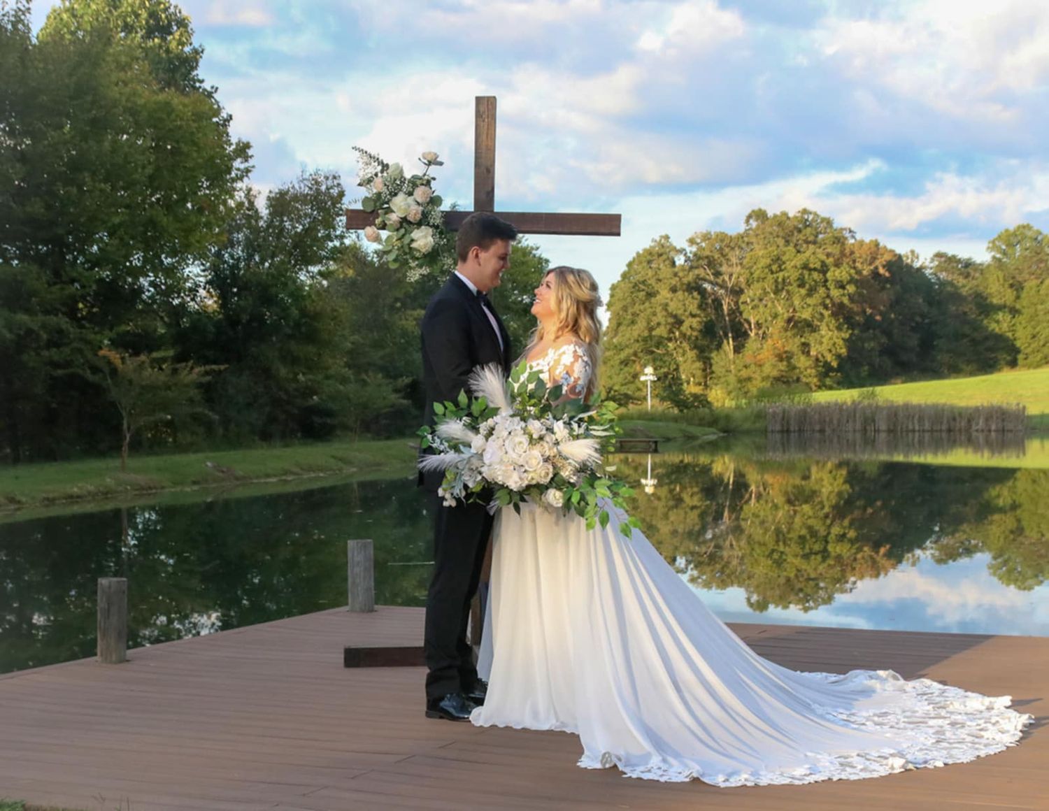 A couple stands on a dock by a lake with a wooden cross, surrounded by natural scenery.
