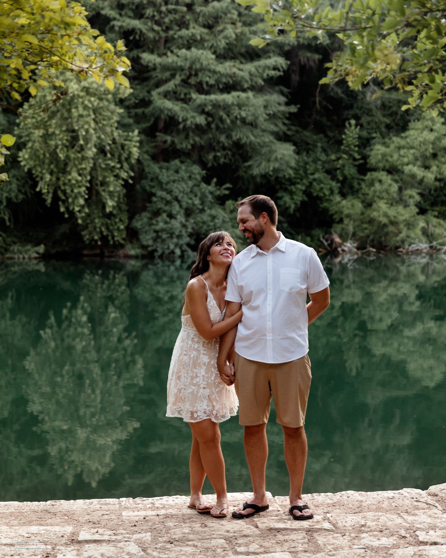 A couple stands together on a stone riverbank at Red Bud Isle, surrounded by green trees and calm turquoise water.
