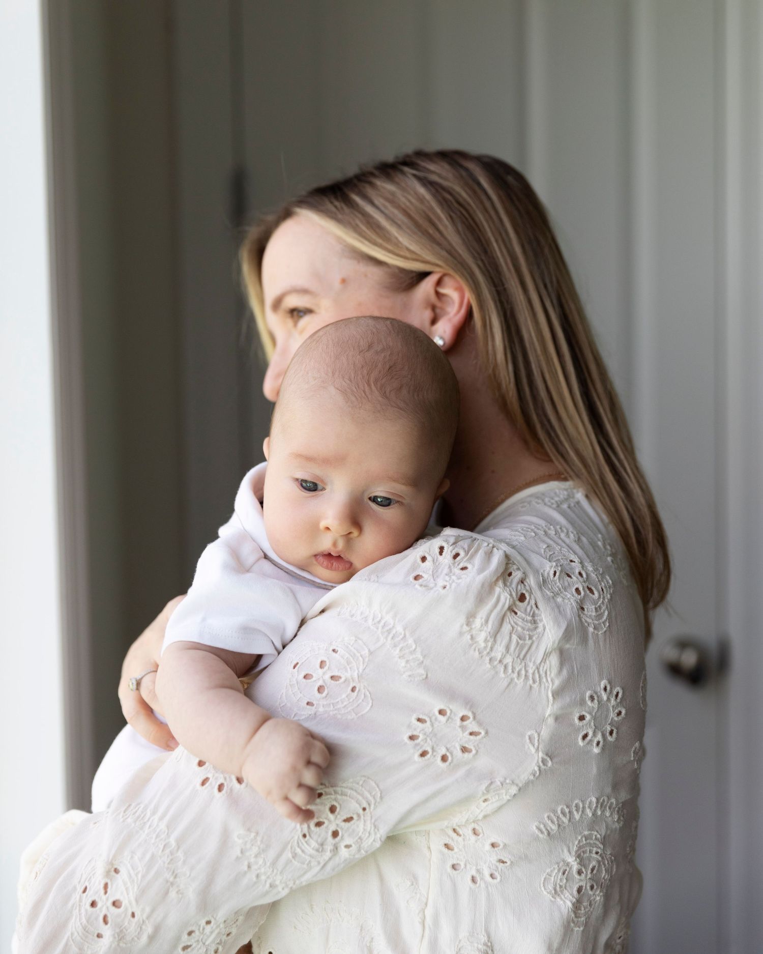 A tender moment by a window as a woman in a white eyelet dress cradles an infant against her shoulder in soft light.