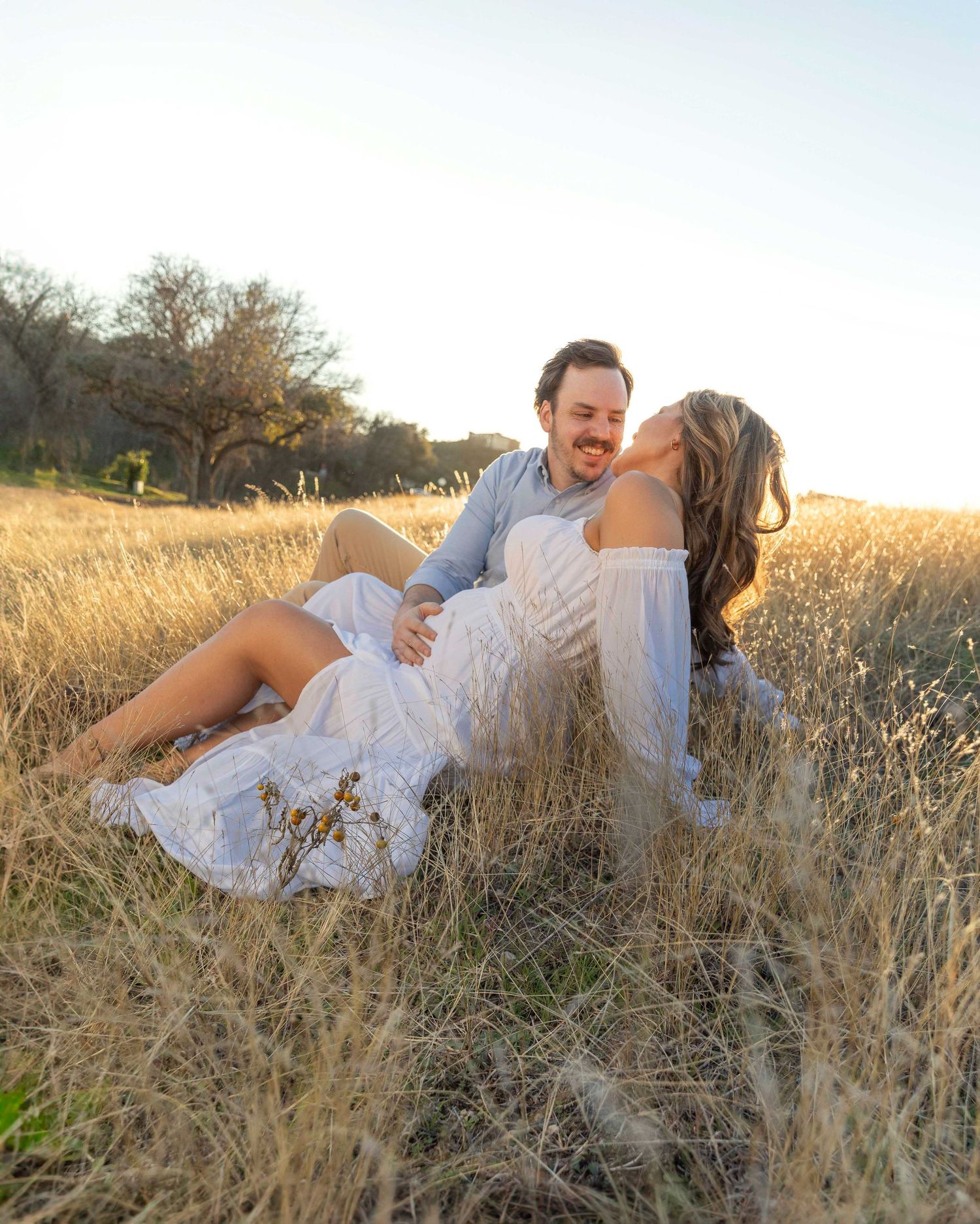 A couple in white clothing sits together intimately in a sun-lit field of golden grass during sunset.