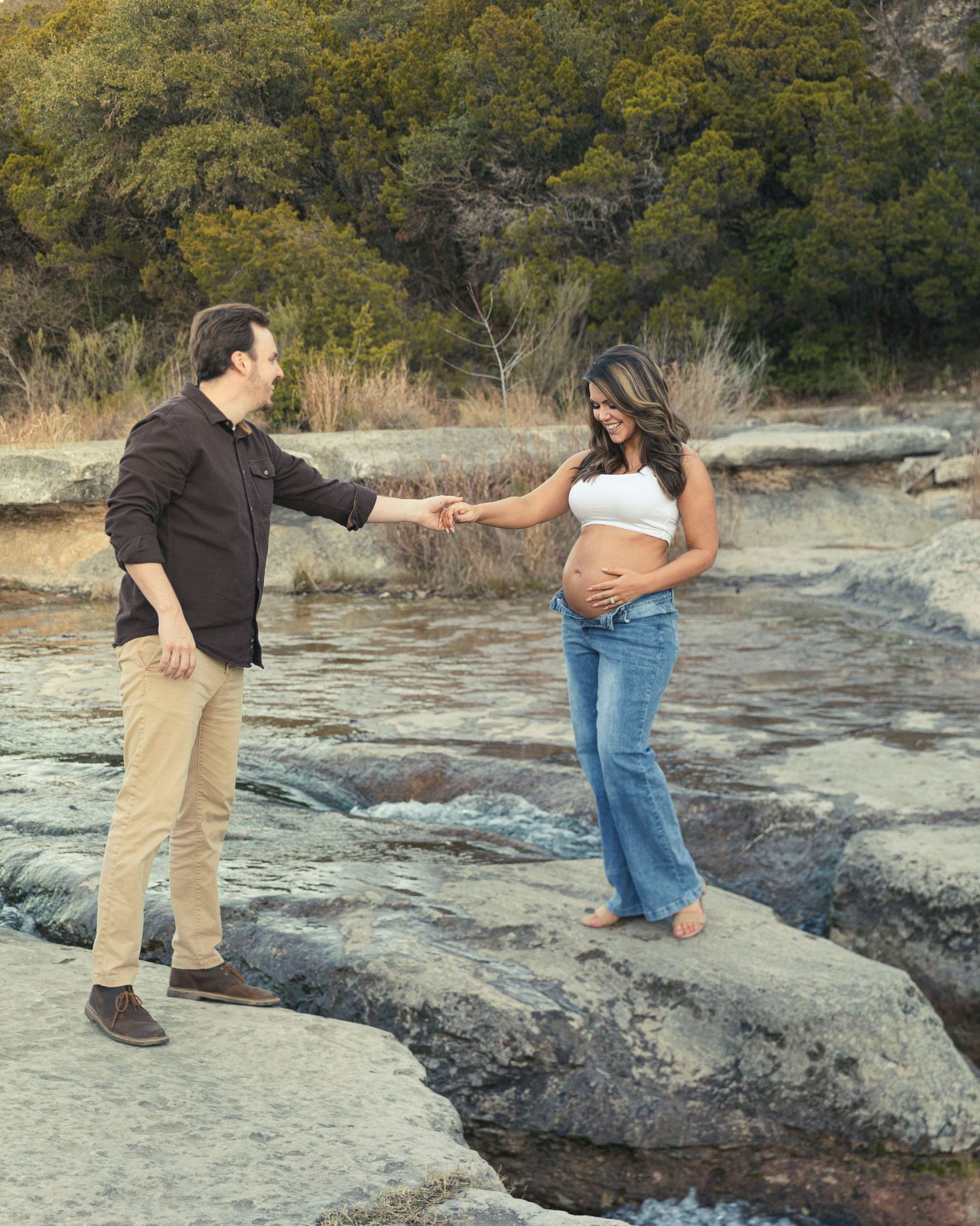 Expectant couple shares a tender moment while standing on rocky terrain near trees during an outdoor maternity photoshoot.