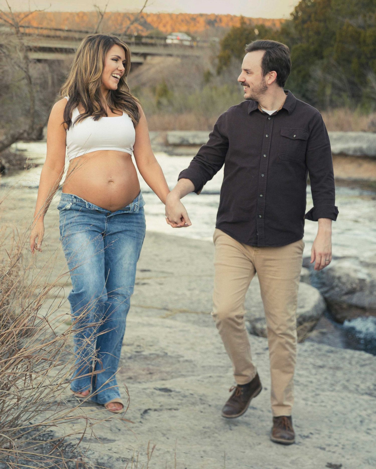 Expectant couple holding hands while walking together on a rocky riverbank during autumn sunset.