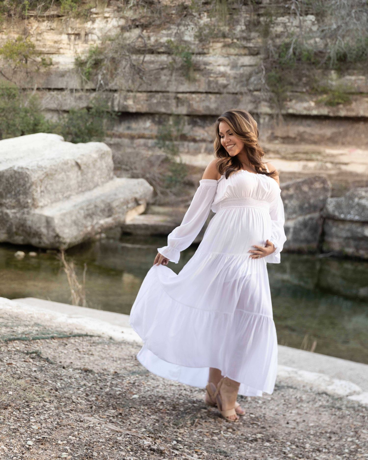 Woman in flowing white off-shoulder dress poses gracefully by natural quarry pool with limestone cliffs in background.