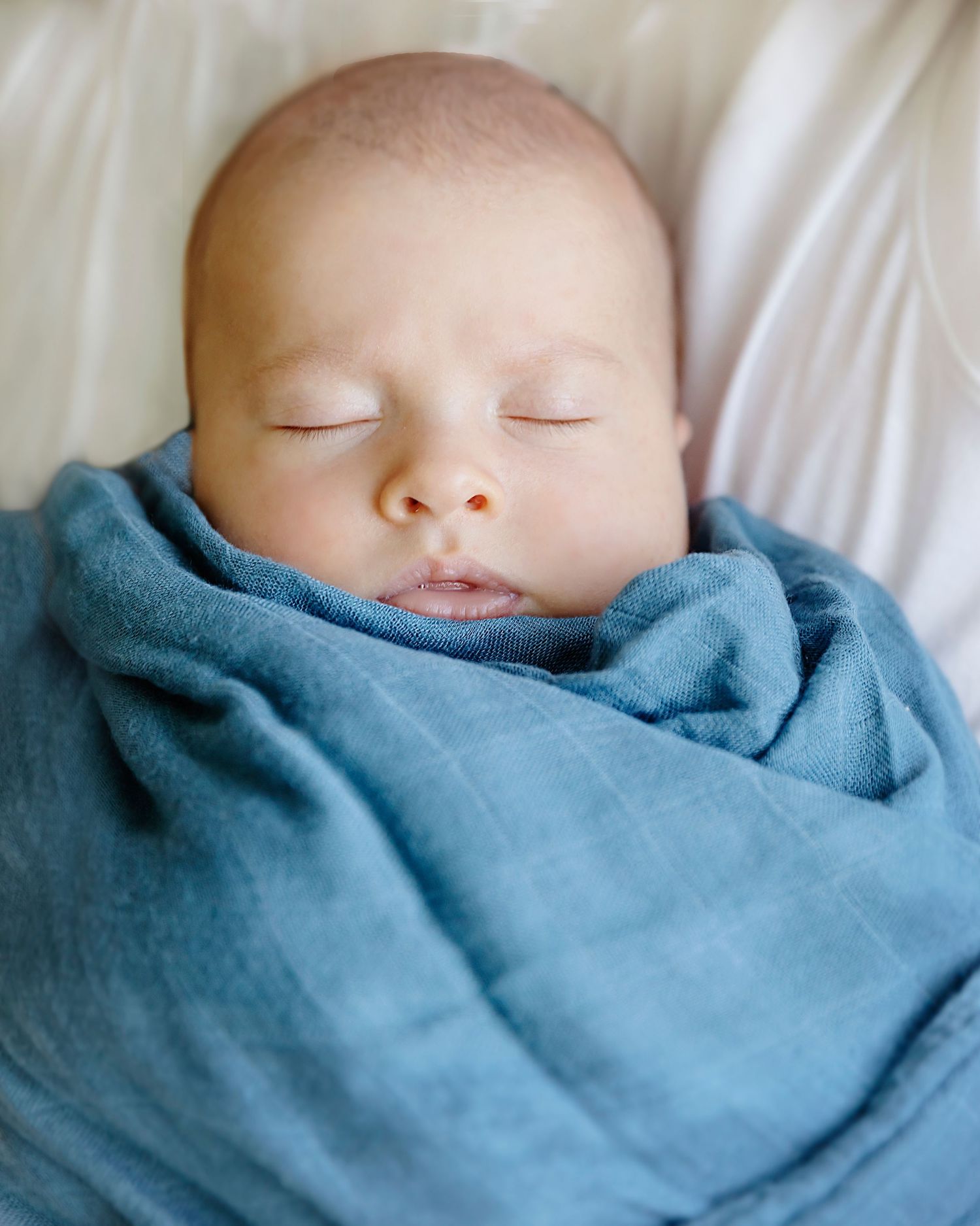 A sleeping infant swaddled in a soft blue blanket rests peacefully against white bedding.