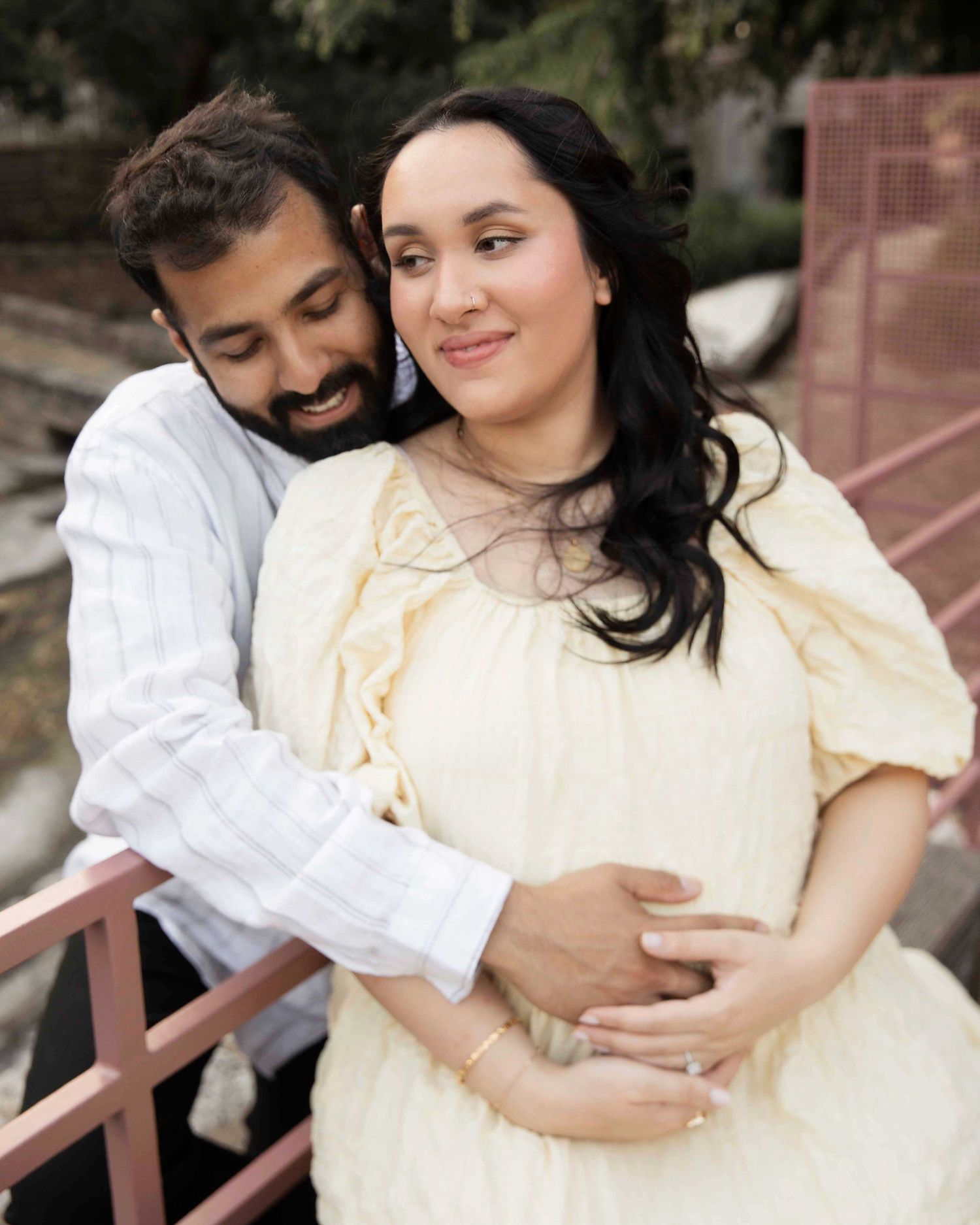A couple embraces outdoors in a romantic maternity photo, with her wearing a cream-colored dress and him in a white shirt.