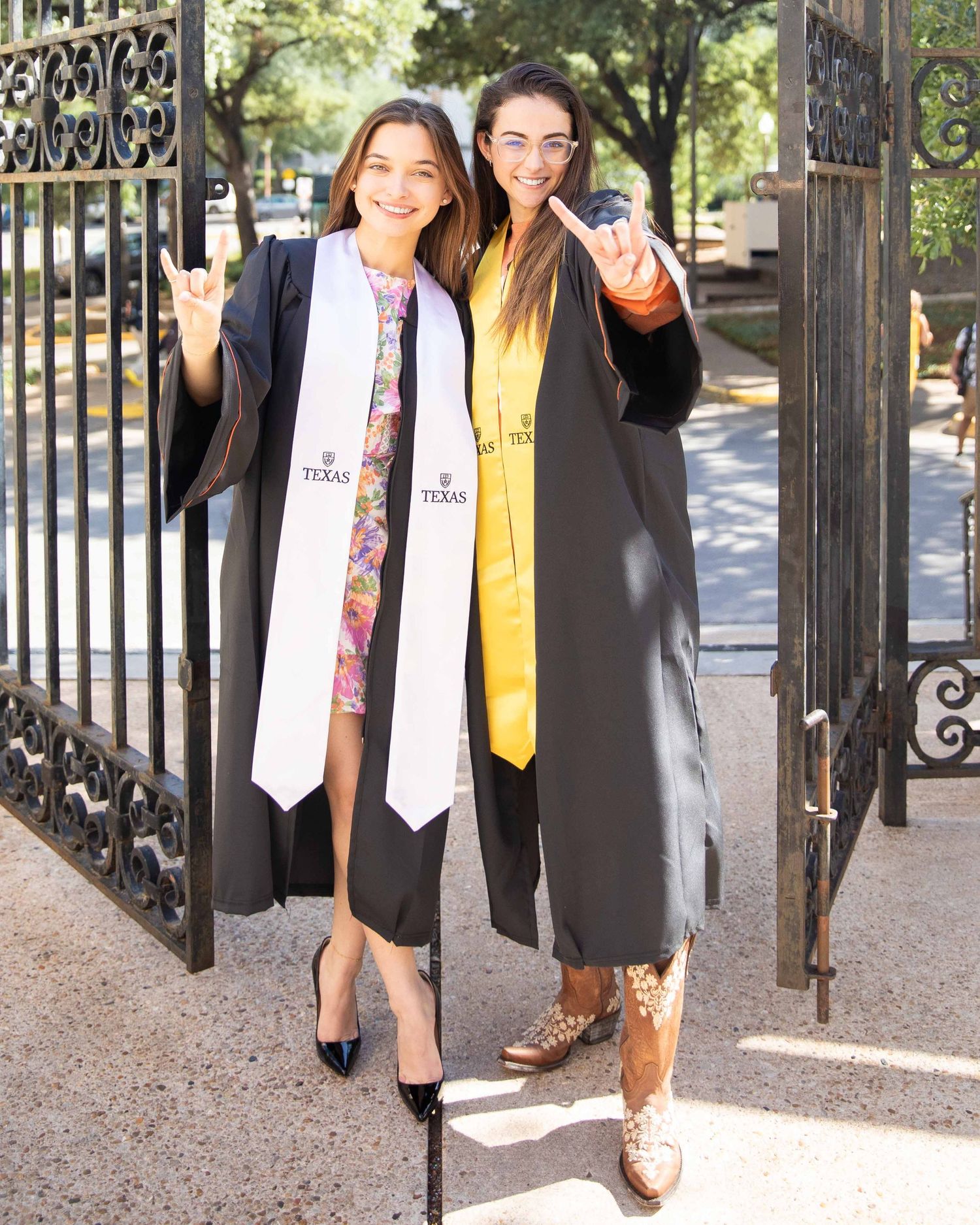 Two graduates in black caps and gowns pose together by ornate metal gates on a UT Austin campus path.