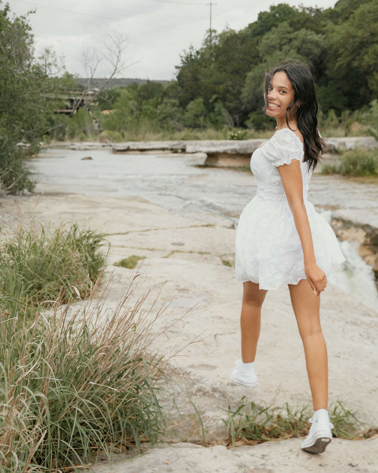 A cute Senior Girl  in a short white lace dress poses on rocky terrain near a river with greenery in the background.