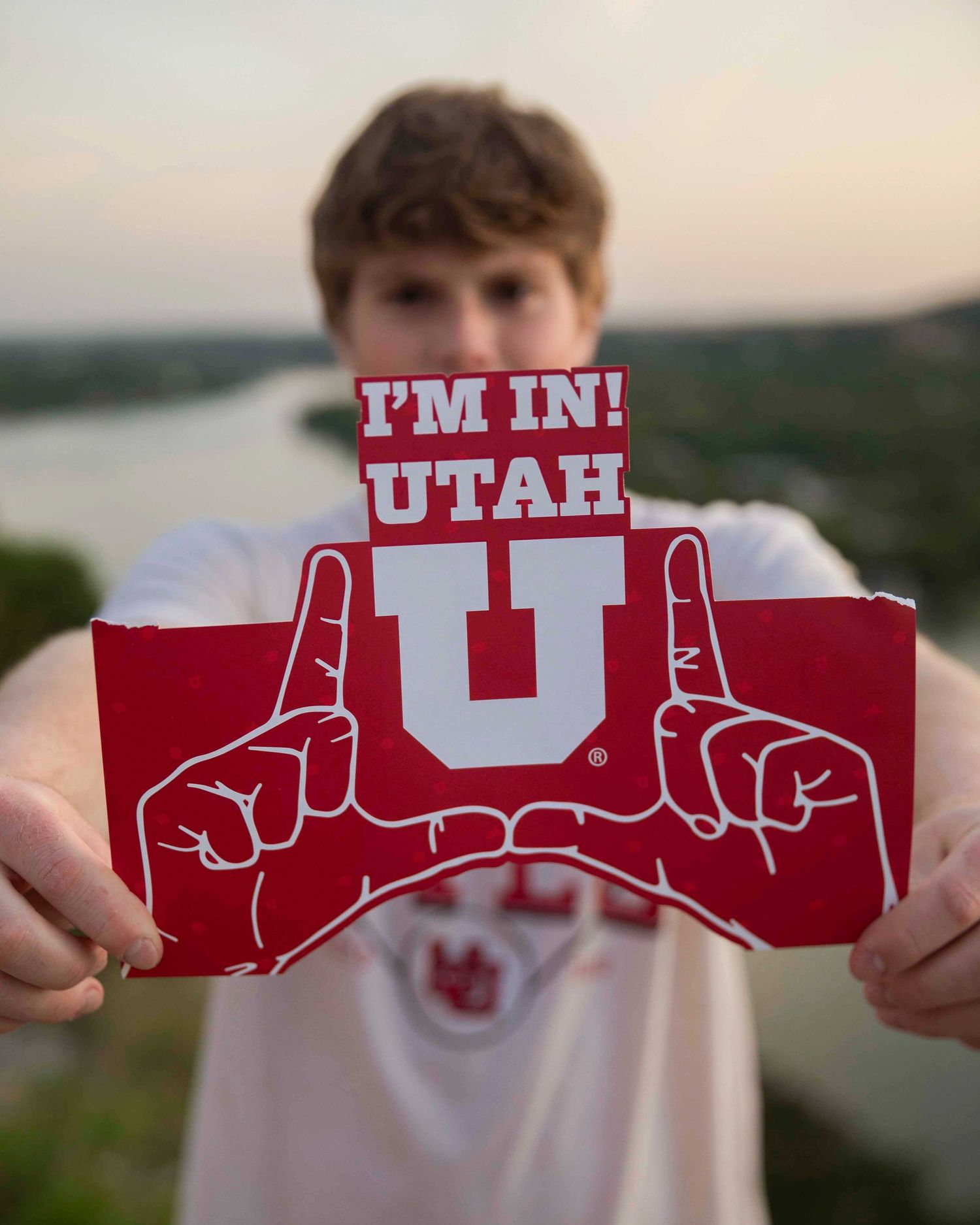 A senior boy up a red University of Utah spirit sign with the U logo and text 'I'M IN!' against a sunset landscape.