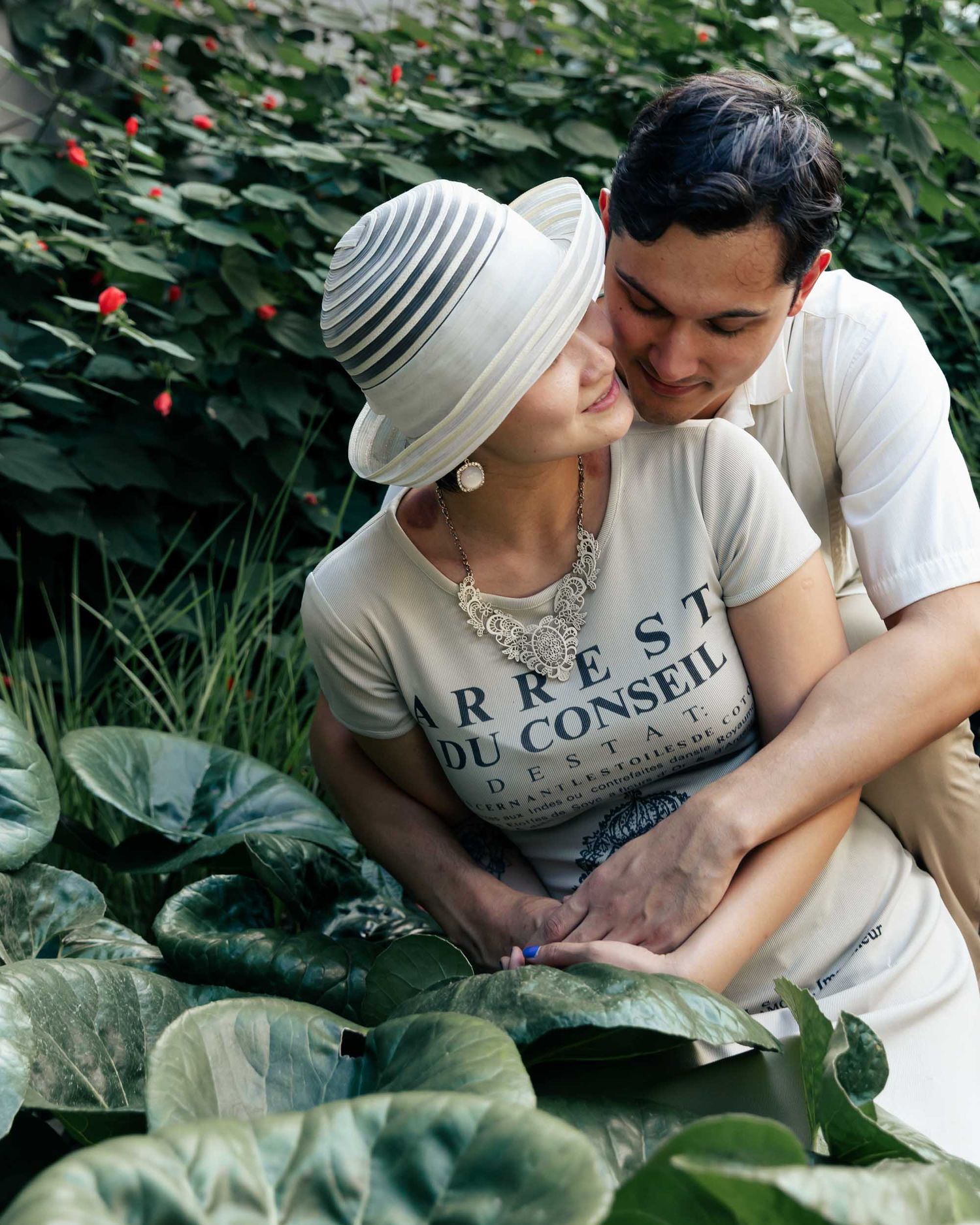 A tender romantic moment shared between a couple in a lush garden setting with green foliage and red flowers in the background.