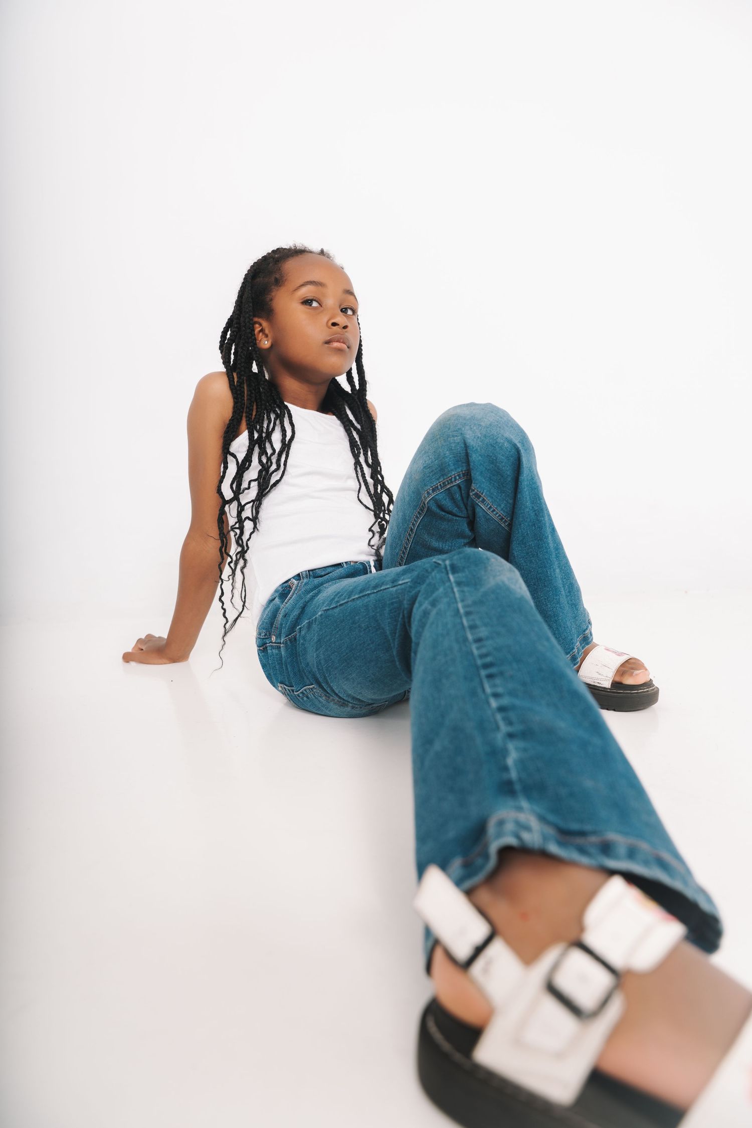 Young Black girl with long braids sitting on white studio floor in jeans.
