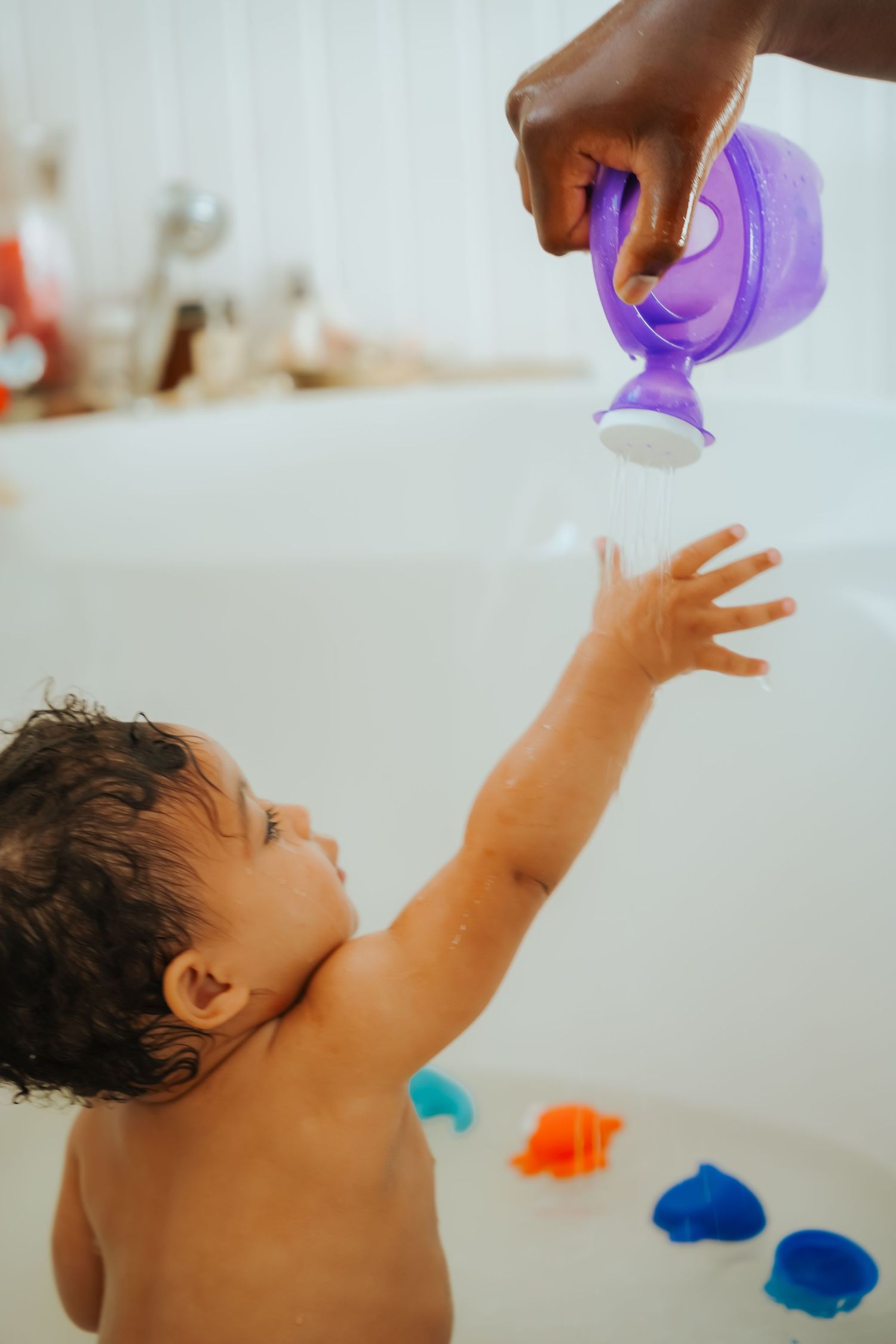 Parent pours water from purple watering can toy over reaching baby during bath time.