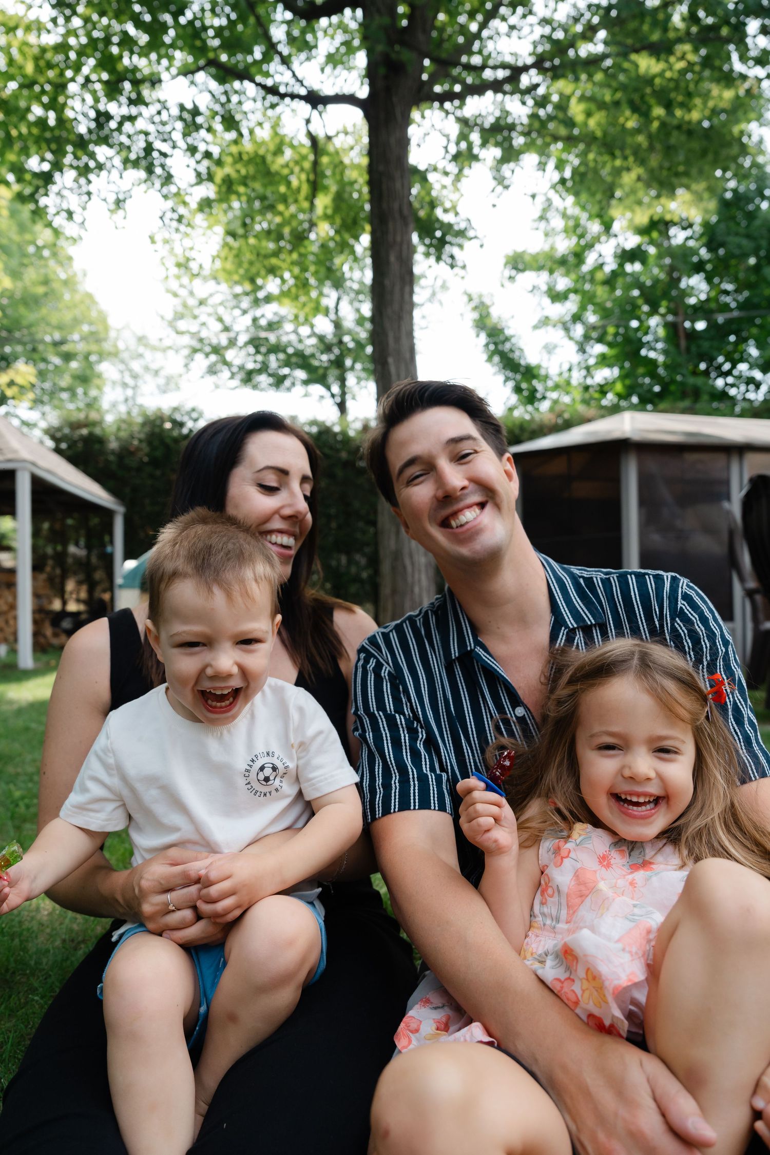 Happy family of four smiling outdoors, two parents holding young children in a lush green backyard setting.