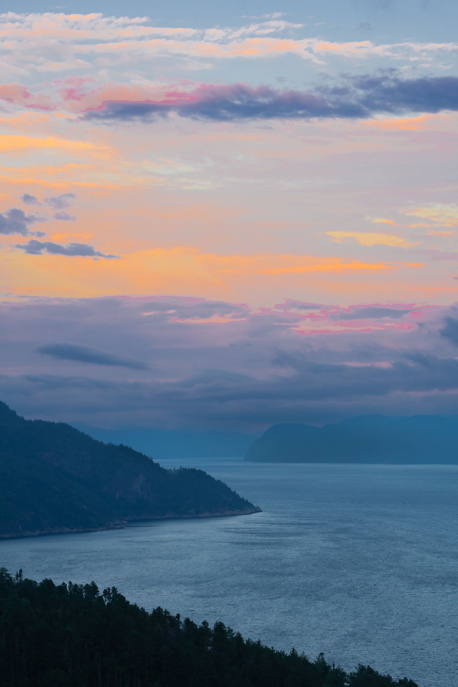 Stunning sunset over a calm lake with colorful pink and orange clouds, misty mountains, and dark forested shoreline.