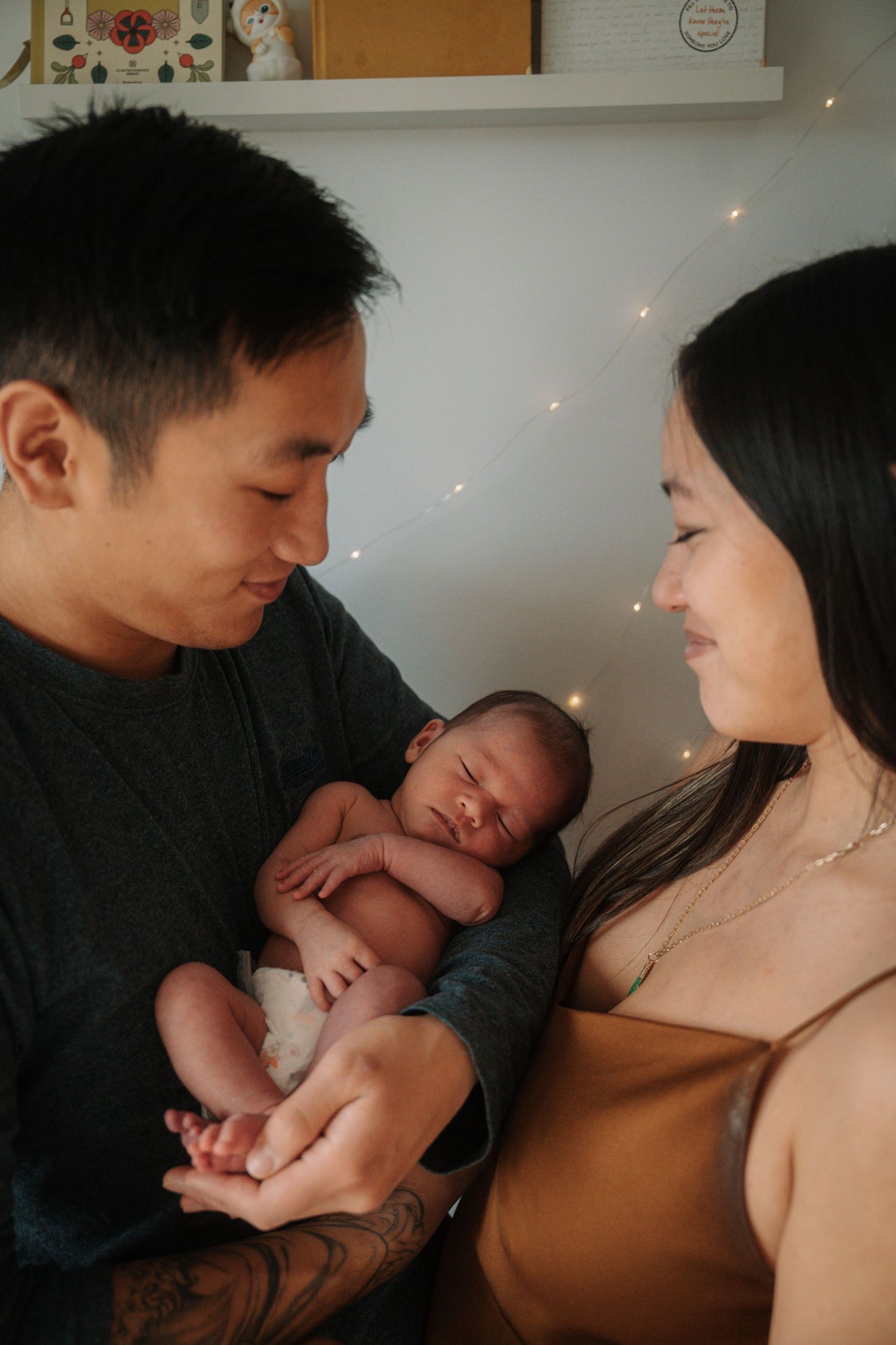 Asian couple lovingly gazing at their sleeping newborn baby held between them with fairy lights behind.