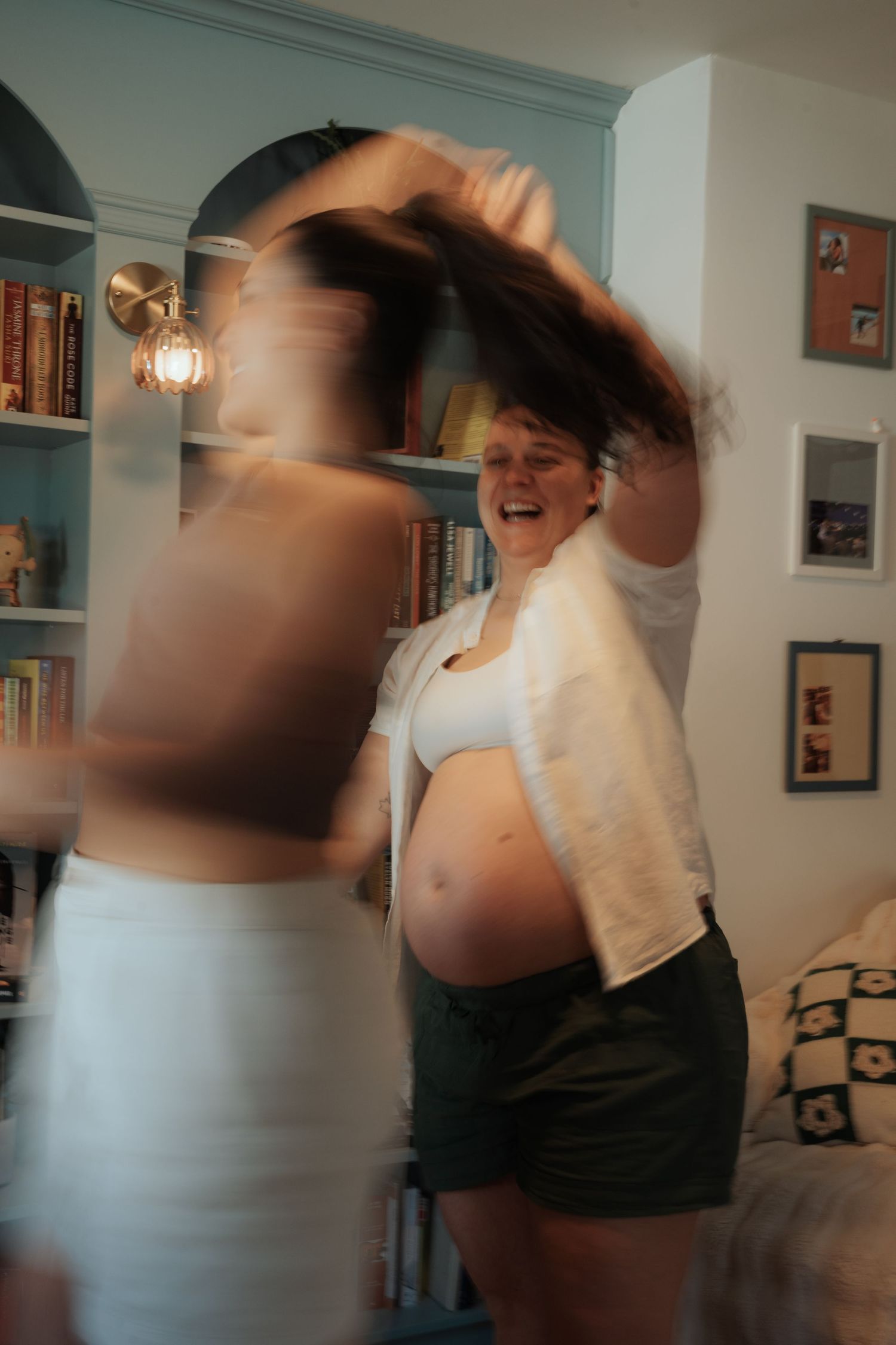 A joyful pregnant woman dancing with a friend in a warm, book-filled home interior with soft lighting.