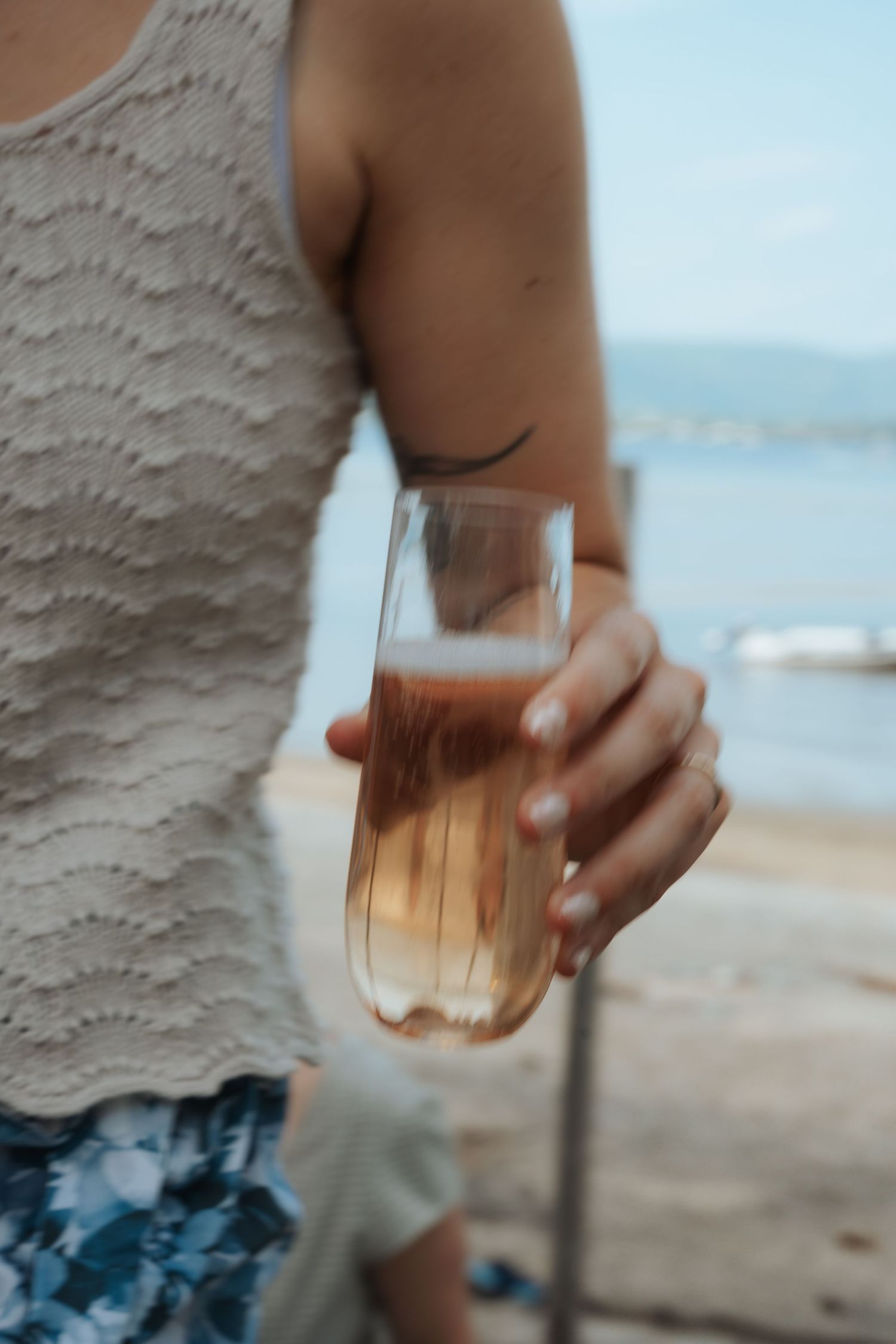 A person in a white lace tank top holds a champagne flute with rose wine near a lakeside beach.
