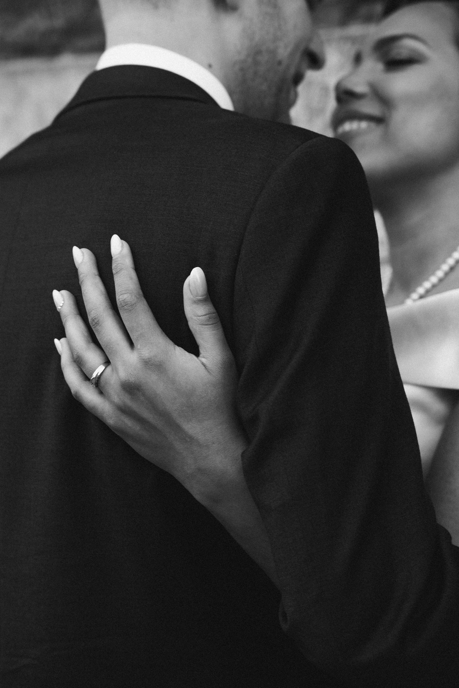Black and white close-up of a bride placing her hand on the groom's back, wedding ring visible.