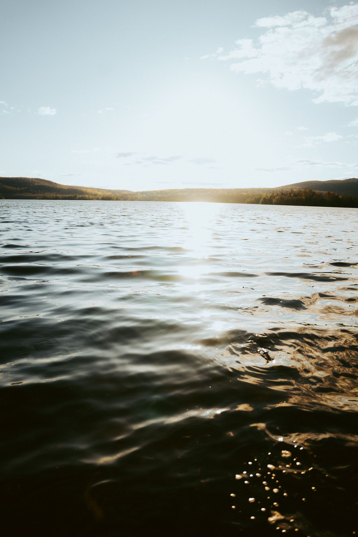Golden sunset reflecting on calm lake waters with rolling hills and a partly cloudy sky in the background.