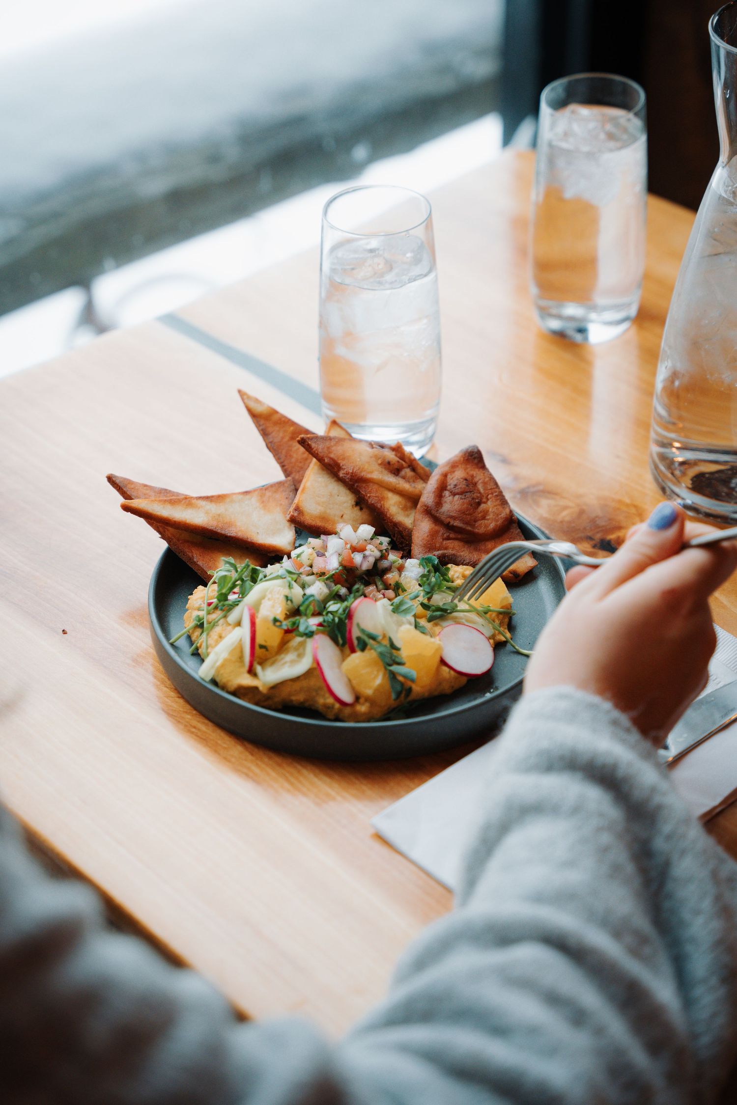 Person eating fresh salad with radishes and crispy flatbread on dark plate at bright restaurant window.