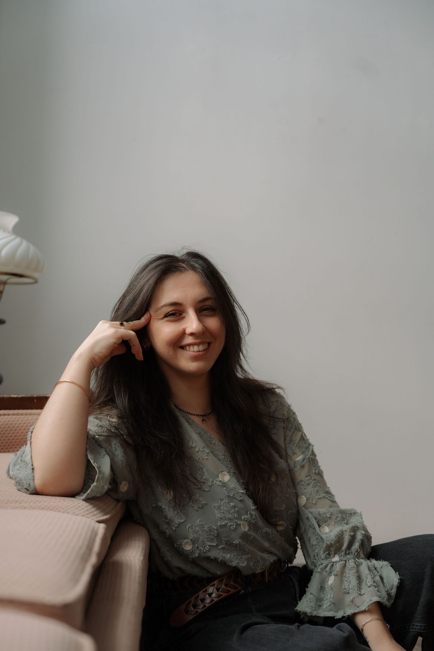 Smiling young woman with long dark hair in green floral blouse relaxing on beige sofa.