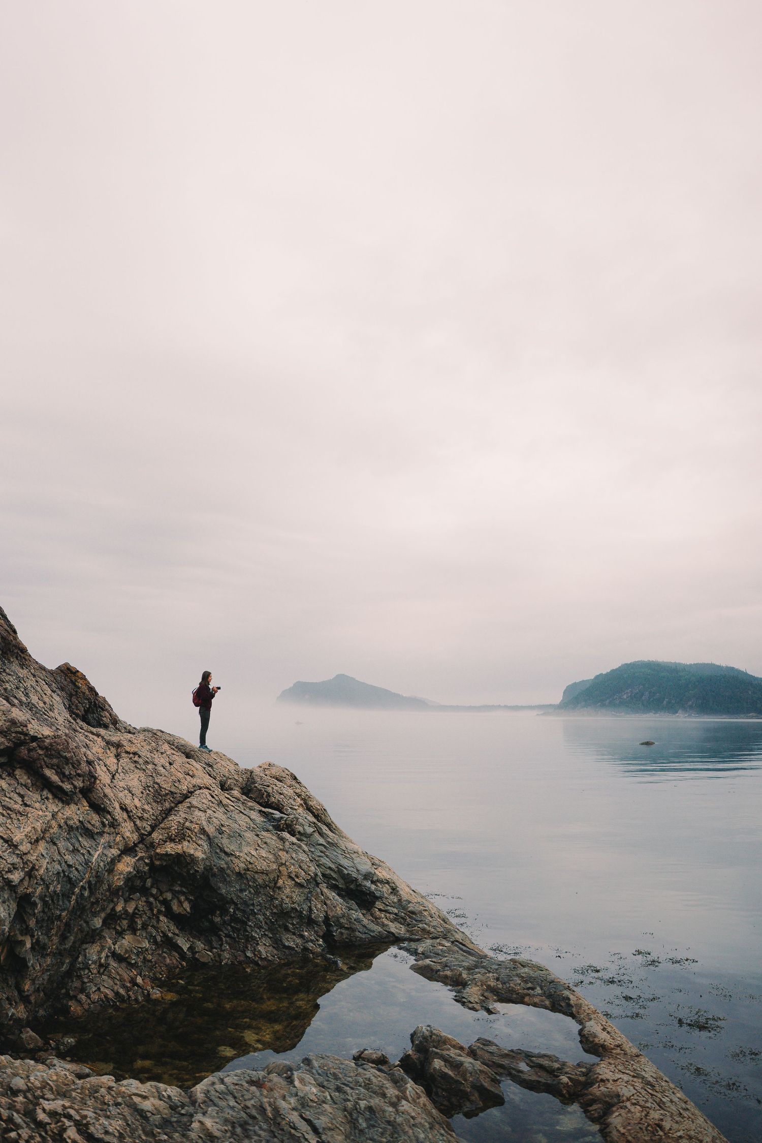 A lone hiker stands on rocky coastal cliffs overlooking a calm misty sea with distant islands under an overcast sky.