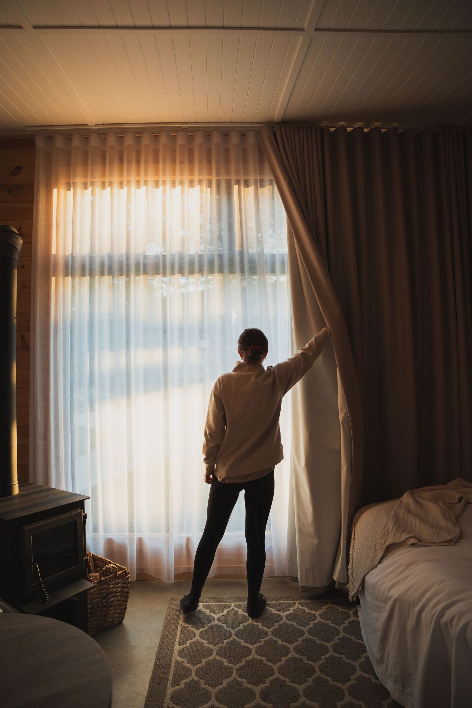 Woman opening sheer curtains to let sunlight into rustic bedroom with wood stove.